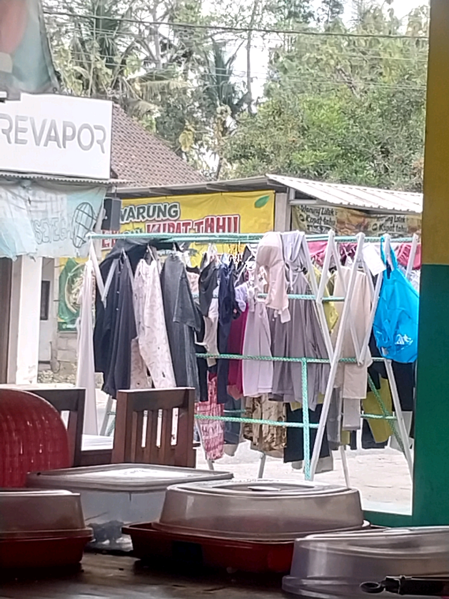 The image captures an outdoor scene in Sayegan, Indonesia, likely a commercial area. In the foreground, multiple food containers with clear plastic lids are arranged on a surface, suggesting a food stall or market setting. Behind these, a clothes rack is set up with various garments hanging on it, including shirts, pants, and a bright blue bag. The rack itself is made of white metal.
In the background, a building with a tiled roof is visible, along with some vegetation. A prominent sign reads "REVAPOR" in large letters, indicating a business. Another sign above the clothes rack features yellow lettering and some text in Indonesian, possibly advertising food items. The overall impression is one of a bustling local establishment where goods, including food and clothing, are displayed for sale in an open-air environment. The lighting suggests it is daytime, with natural light illuminating the scene.