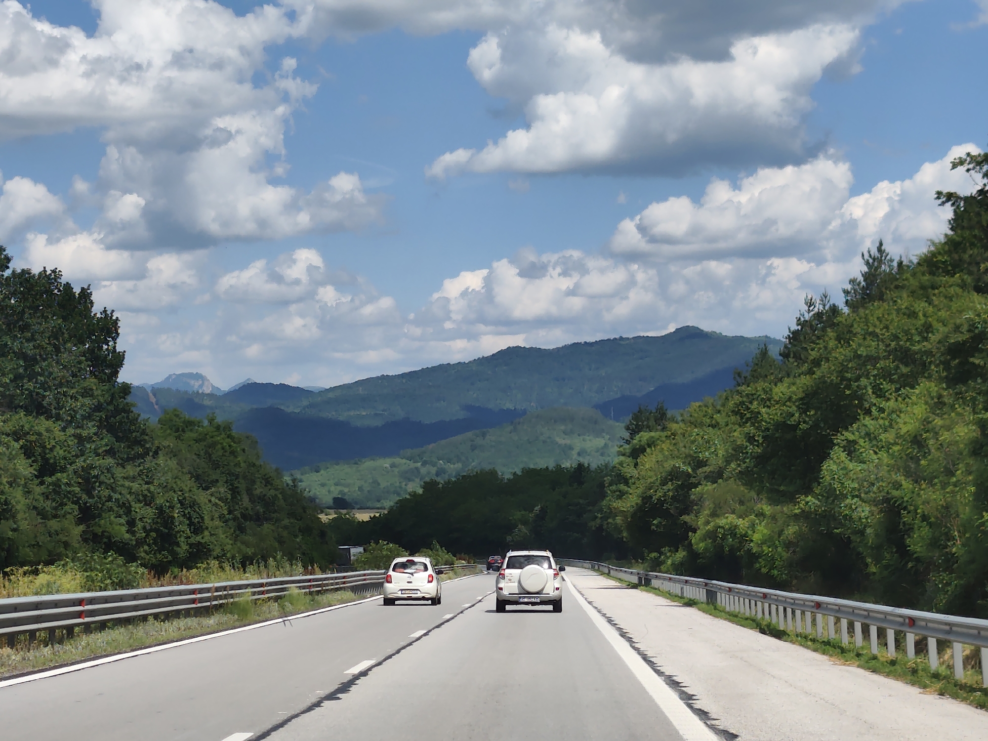 The image depicts a sunny day on a highway with a clear blue sky and fluffy white clouds. The road appears to be a divided highway with two lanes in each direction. Guardrails line both sides of the road. In the foreground, two cars are traveling away from the camera. The car on the left is a white hatchback, and the car on the right is a white SUV with a spare tire mounted on the back. Further down the road, another car can be seen in the distance. The highway is surrounded by lush green trees and rolling hills in the background. The overall scene suggests a rural or mountainous landscape. The location is the highway in Osikovitsa, Bulgaria.