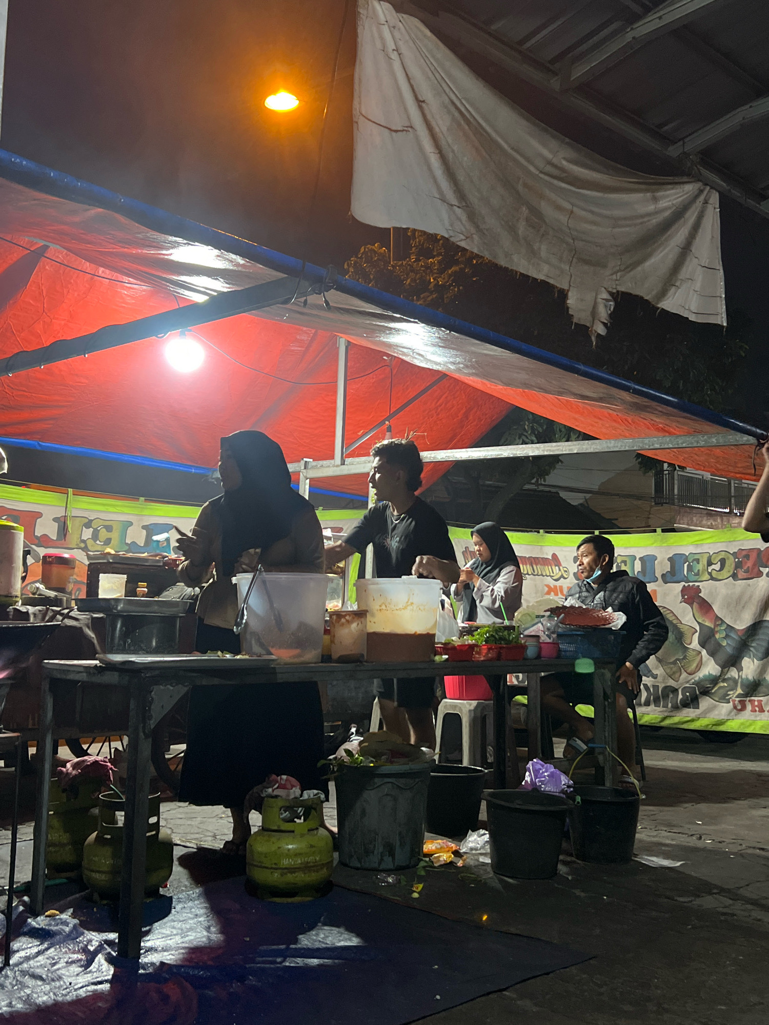 At night in Tlogoadi, Indonesia, a street food vendor stall is set up under a makeshift awning of red and white tarps. A bright overhead light illuminates the scene. Three people are visible: a woman in a headscarf and brown jacket is serving food, a man in a black t-shirt is preparing food, and another man in a black jacket and face mask is seated at a table. Several propane tanks are on the ground near the stall, along with buckets and assorted refuse. A banner with colorful graphics of roosters and fish is displayed in the background. The overall atmosphere suggests a bustling evening at a local eatery.