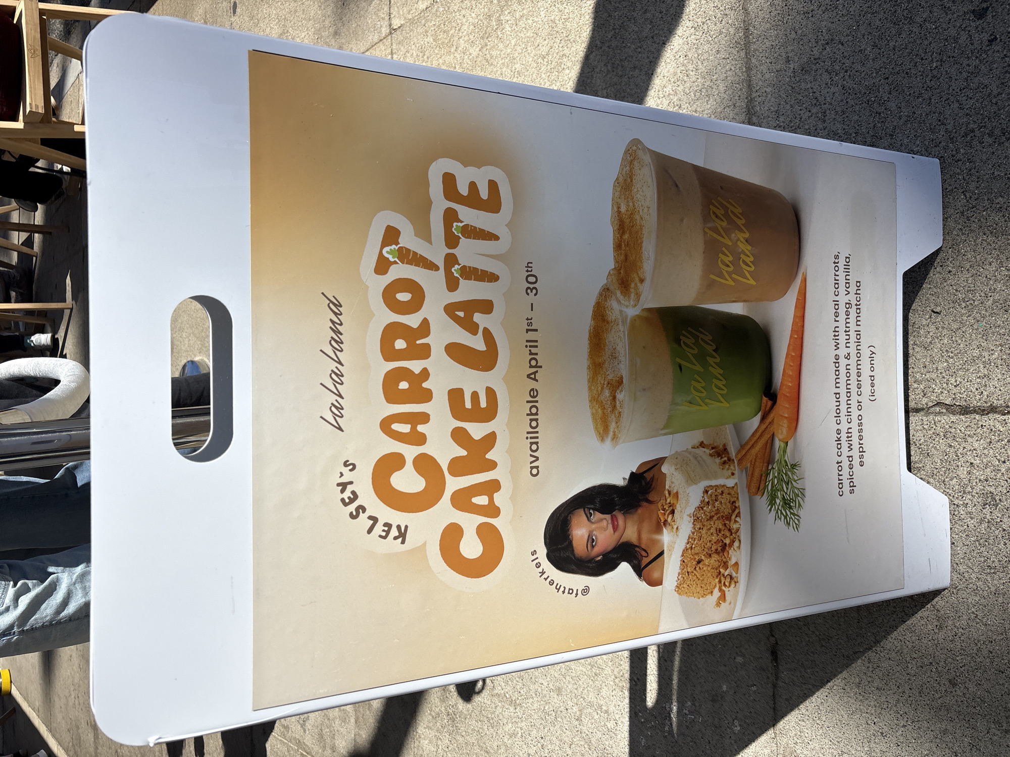 The image shows an advertisement for a "Carrot Cake Latte" by Kelsey's Lalaland, available during April 1st-30th. The advert, printed on a sandwich board, is placed on a sidewalk, likely in a city environment. The sign has a white frame with a cutout handle at the top. The advert has a beige background. The advert features photos of the drink in cups, as well as an image of a carrot cake. The advert also has the Instagram handle @fatherkels. The drink is described as a "carrot cake cloud made with real carrots, spiced with cinnamon & nutmeg, vanilla, espresso or ceremonial matcha (iced only)." The location is likely in Newport Beach, United States of America.