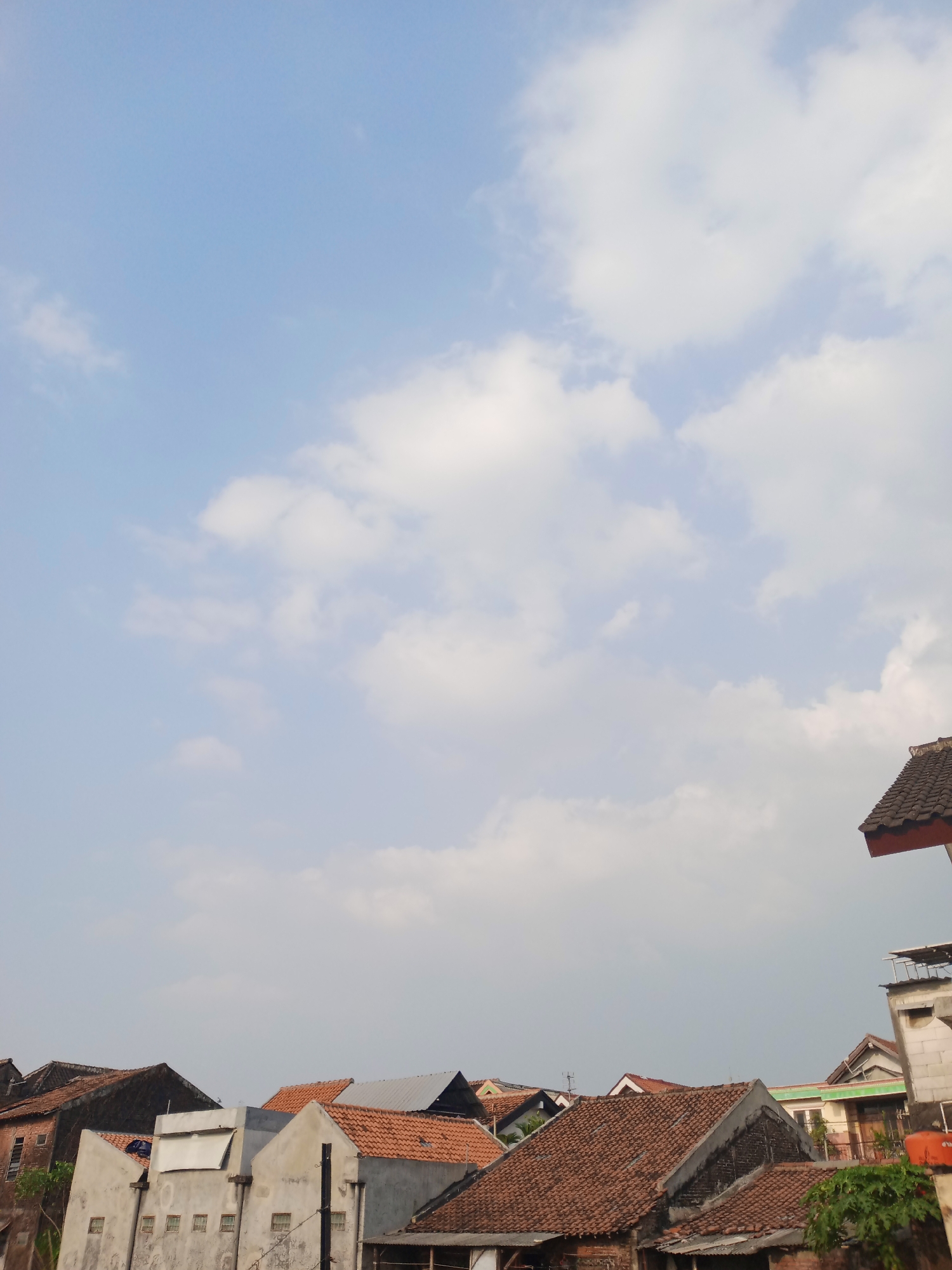 The image depicts a view of rooftops and the sky in what appears to be a residential area. The architecture suggests a tropical or sub-tropical climate, with many buildings featuring terracotta tiled roofs. The sky is a pale blue with scattered, wispy white clouds. The overall impression is one of an urban or semi-urban environment, possibly in a developing country, given the apparent age and style of some of the buildings. While specific details like people or notable landmarks are absent, the image captures a common scene of everyday life, focused on the built environment and the natural sky. The scene could be in Demak, Indonesia, a region known for its traditional architecture and close proximity to the sea, which would explain the atmospheric conditions and building styles.