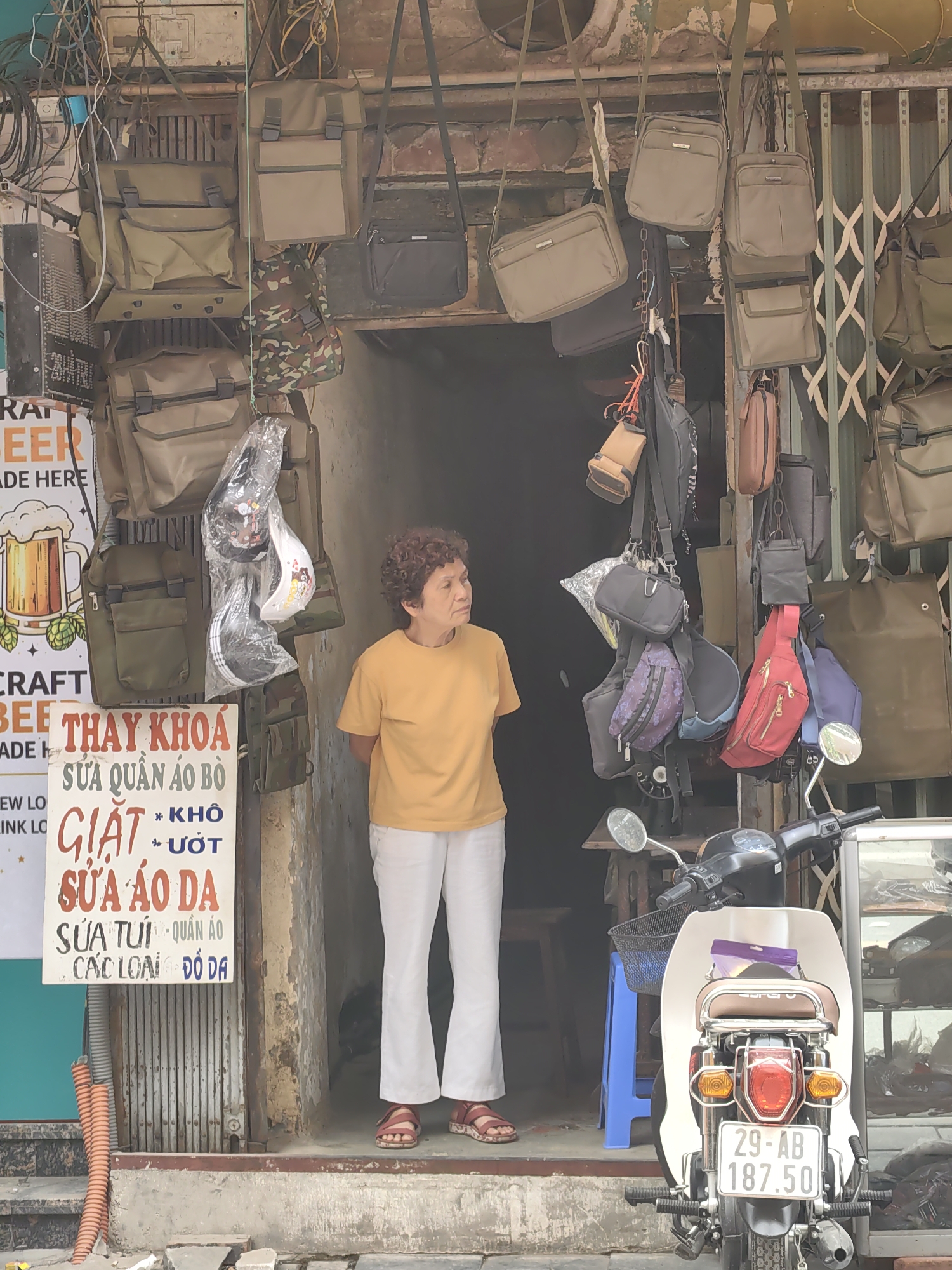 The image captures a woman standing in the doorway of a small shop in what is likely Hà Nội, Vietnam, based on the license plate on the motorcycle. The shop's entrance is surrounded by an array of hanging bags of various styles and colors. A sign on the left advertises tailoring and laundry services in Vietnamese. A white motorbike is parked in front of the shop, partially blocking the view of a blue plastic stool. The woman in the doorway is wearing a yellow t-shirt, white pants, and red sandals, and she is gazing off to the side. The environment suggests a bustling, urban setting with a focus on small-scale retail activity.