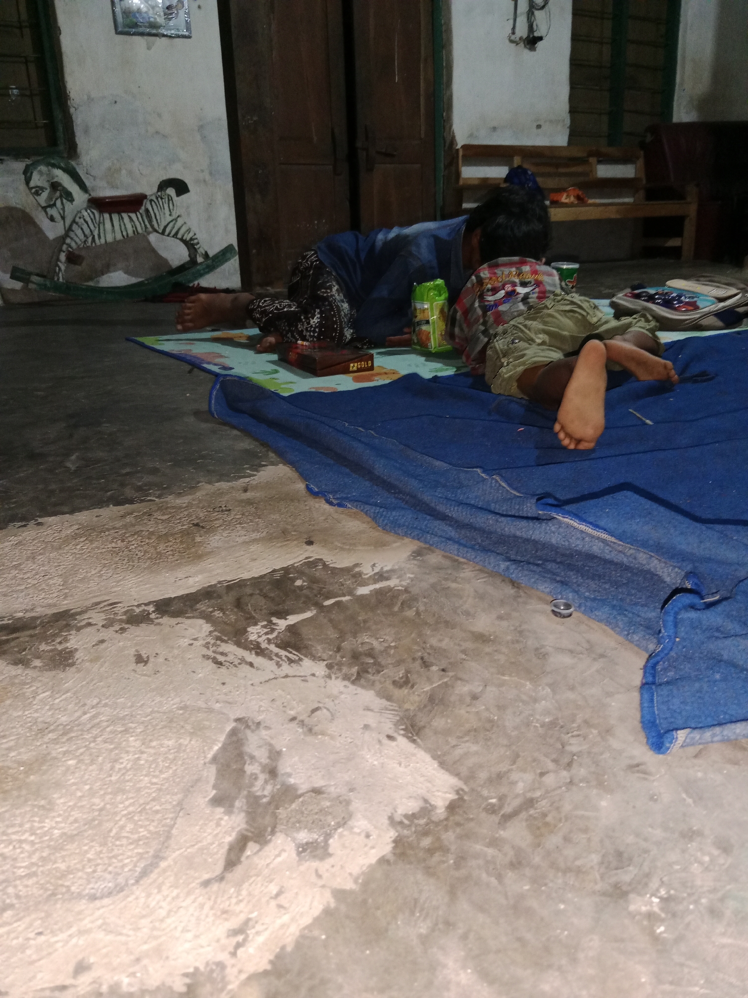 In Ringinrejo, Indonesia, two individuals, likely children, are lying on a blue tarp spread on a concrete floor. The individual in the foreground, wearing green shorts and a plaid shirt, is lying on their stomach with their legs extended, their right foot visible. Behind them, another individual in a dark blue shirt and patterned pants is also lying down. They appear to be interacting with objects on a colorful mat placed between them. On the mat are several items, including a green and yellow food packaging, a dark box with "GOLD" written on it, and a bag. The background shows a wall with a painting of a zebra-like rocking horse and a wooden door. The environment suggests an indoor setting, possibly a home, with simple furnishings. The lighting is dim, casting shadows on the floor. The concrete floor shows patches of wear and discoloration.