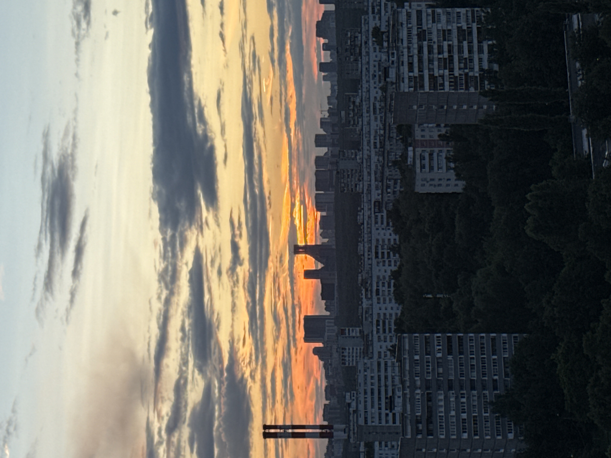 The image shows a skyline of Moscow, Russia, during sunset. The sky is filled with dramatic clouds tinged with orange and yellow hues from the setting sun. Silhouetted against the sky are several buildings, including apartment blocks and industrial chimneys. In the foreground, there are multi-story apartment buildings, characteristic of Soviet-era architecture, with many windows. Dense green trees fill the space between the buildings and in front of the further distant structures. The overall impression is of a densely populated urban environment under a colorful sky.