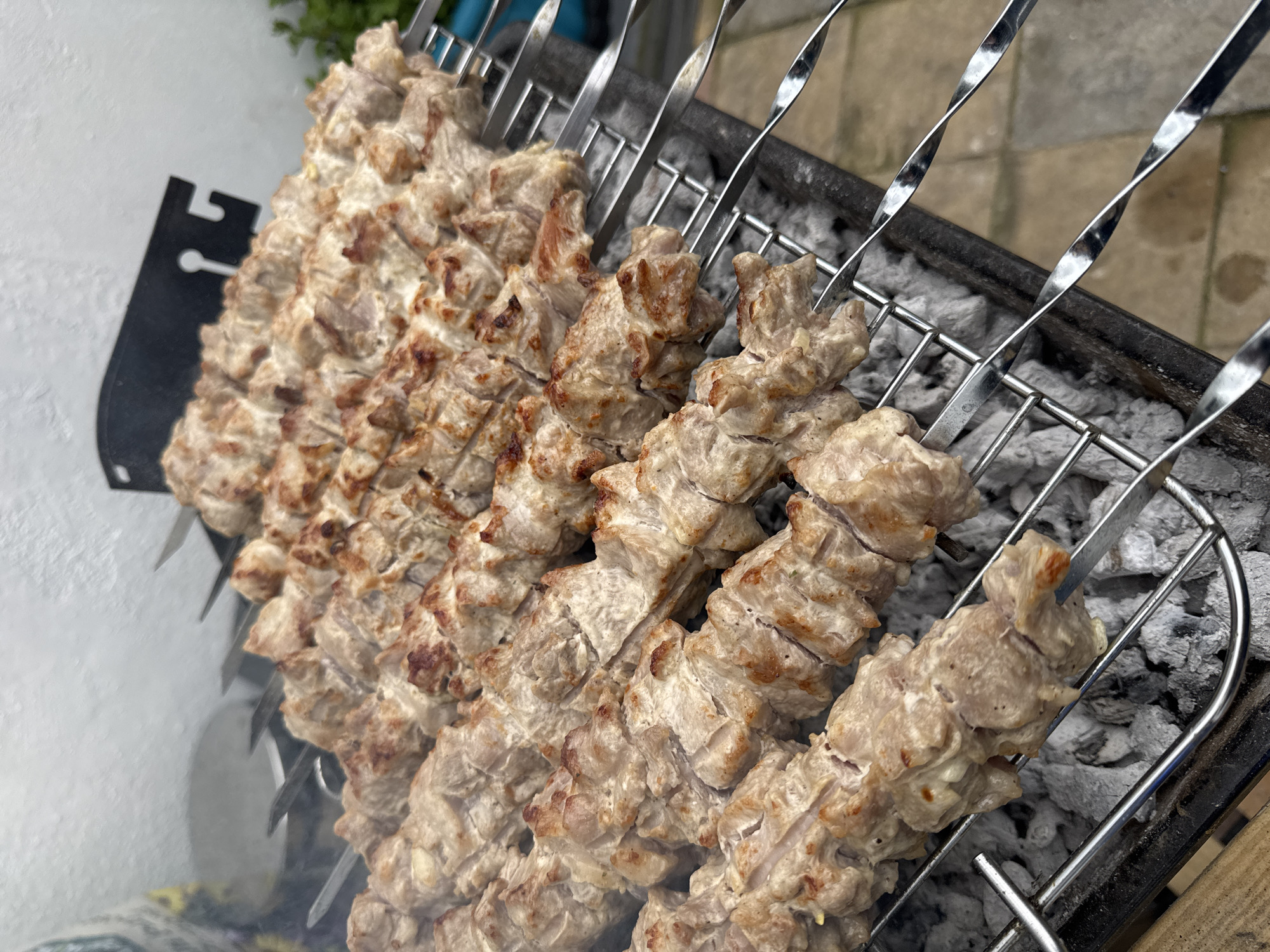 A high-angle shot depicts several metal skewers loaded with what appears to be grilled meat, resting on a charcoal grill. The charcoal underneath the meat is grey and ashy, indicating previous use. Wisps of smoke rise from beneath the food. The skewers have a twisted, spiraling design. The background shows what might be a wall or surface, as well as some greenery. The image doesn't provide sufficient information to confirm its location within the city of Gorleston-on-Sea, United Kingdom. There are no people visible in the frame. The primary action is the cooking/grilling process implied by the placement of the meat on the hot grill.