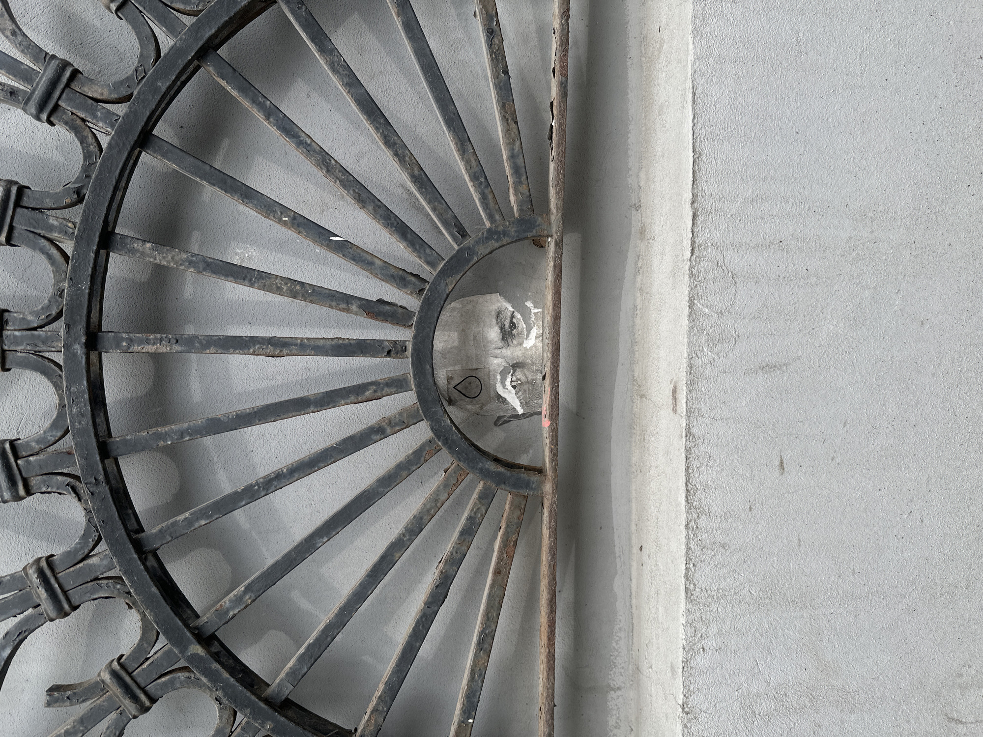 A partially obscured face is visible through a circular metal grate, likely a portion of a fence or decorative architectural element, mounted on a grey concrete wall. The grate's design resembles a sunburst pattern, with decorative flourishes on the outer ring. Flaking paint and rust are visible on the metal. A torn sticker or piece of paper is affixed above the face. The photograph appears to be taken in Moscow, Russia, based on architectural details and the style of the metalwork.