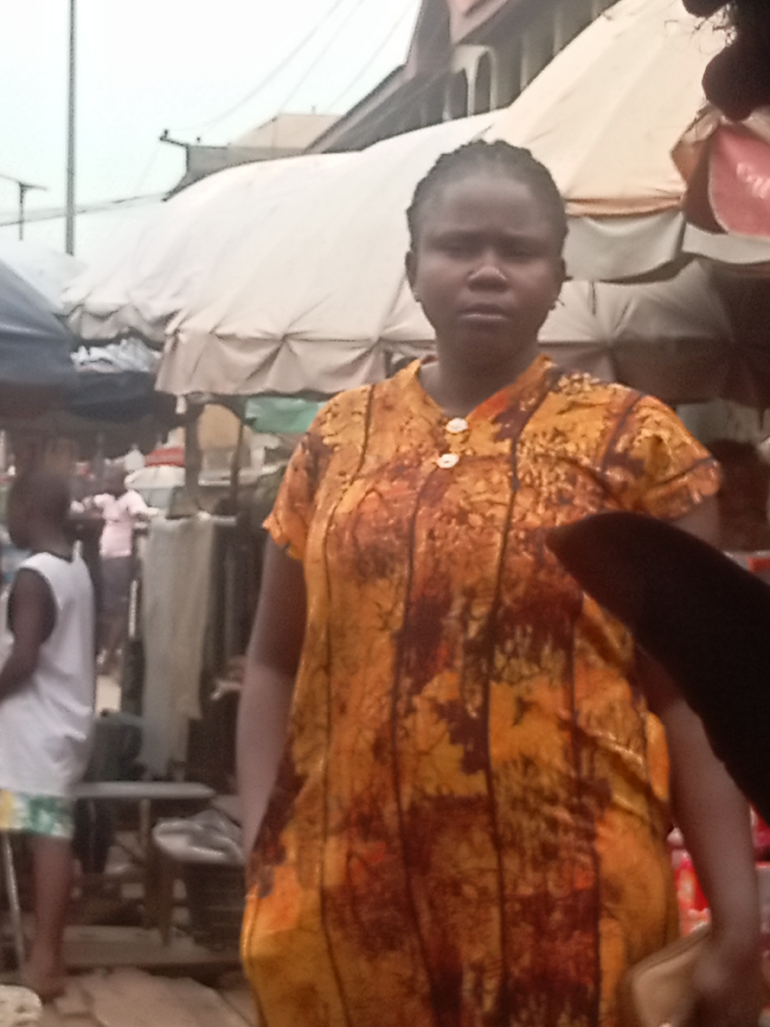 The photo shows a woman standing in a market in Nkpor, Nigeria. The woman is wearing a yellow and brown patterned dress with two white buttons at the neck. She has braided hair. The market stalls in the background are covered with white tarps. Other people are visible in the background, including a person wearing a white tank top and shorts.