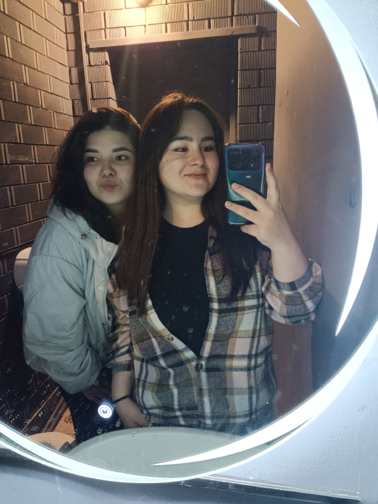 Two women are posing for a selfie in a bathroom mirror with a decorative light border. The woman on the left is wearing a light grey puffy jacket and has dark hair. The woman on the right is holding a teal smartphone to take the picture. She is wearing a black shirt under a plaid button-down shirt. The bathroom features brick-like tiles on the walls and a modern sink. The city and exact location are not discernible from the image.