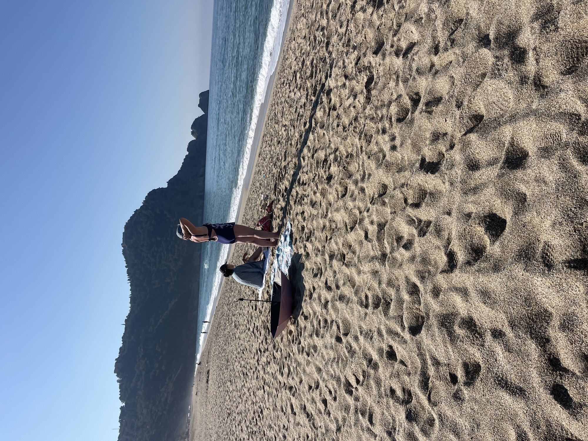 The image shows a beach scene in Alanya, Turkey. A woman in a bathing suit stands on a towel laid out on the sand, near the shoreline. Another person sits nearby. An umbrella is also visible. The sand is textured, with visible footprints and patterns. In the background, there's a mountain range, and the sea meets the shoreline with gentle waves. The sky is clear blue.