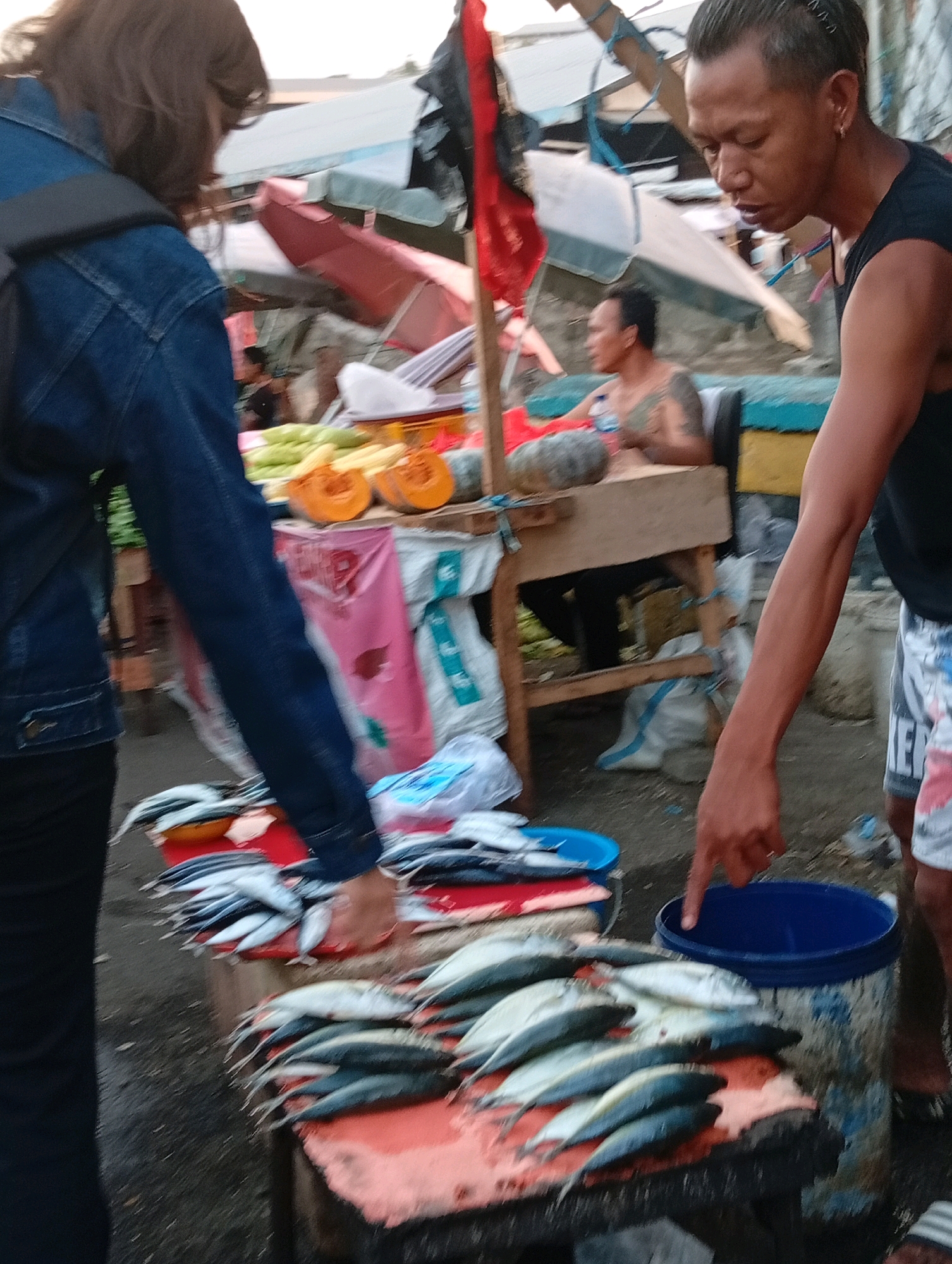 The image depicts a street market scene in Manado, Indonesia. In the foreground, a person wearing a denim jacket and black pants is visible from the back, facing a stall. In the mid-ground, another person, a man with short hair and wearing a black sleeveless shirt and patterned shorts, is pointing at a blue bucket. He is standing near a stall displaying various items, including fish, pumpkins, and corn. In the background, more stalls and people are present, suggesting a busy marketplace. The overall environment appears to be outdoors, with evidence of a somewhat informal setup typical of local markets.