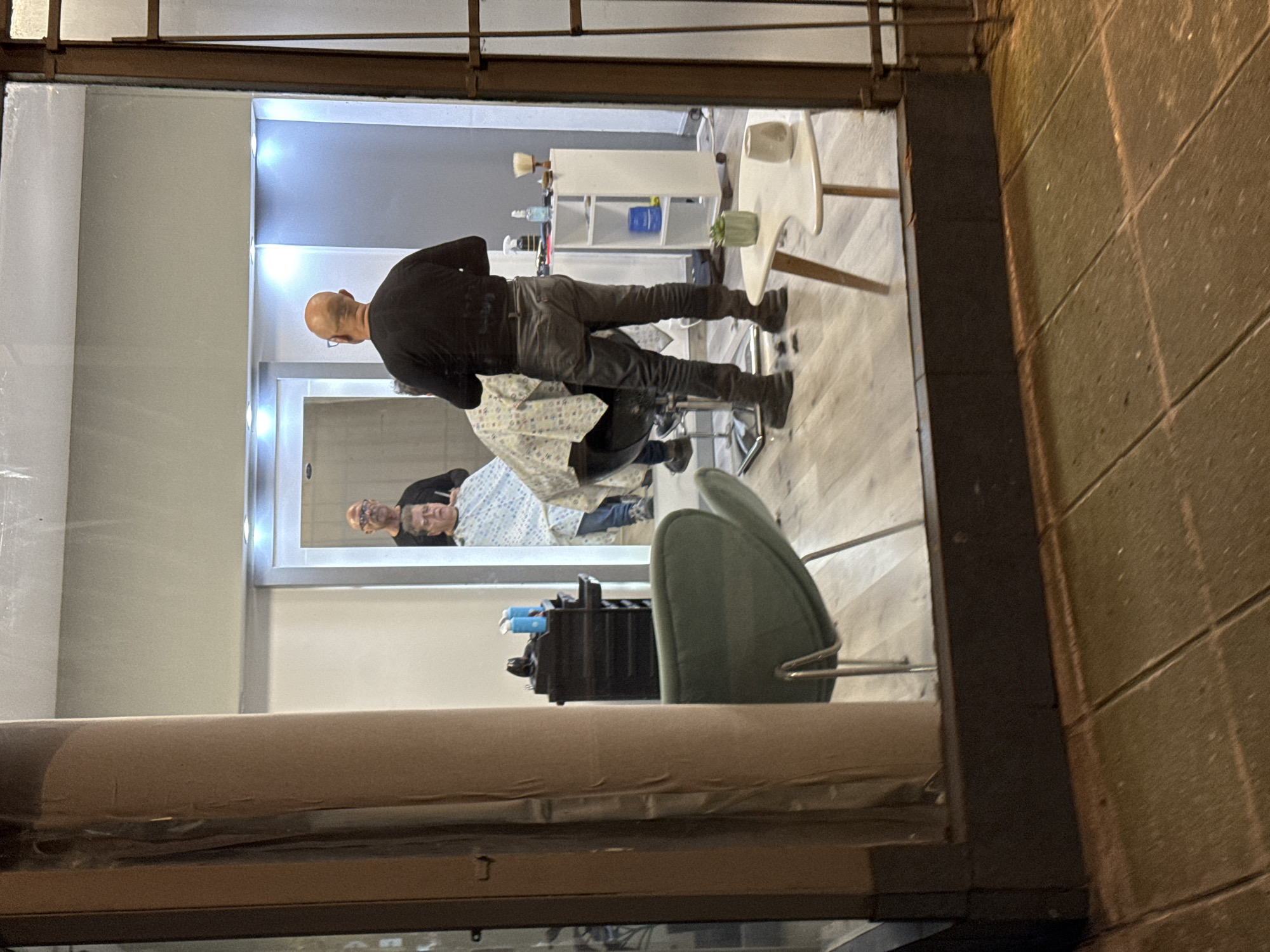 The image captures the interior of a hair salon, likely at night, as seen through a window with a dark frame. The reflection of a street scene or a building wall is visible on the window's surface.
Inside, a bald man wearing a black long-sleeved shirt and gray jeans is standing and appearing to be cutting the hair of a seated customer. The customer is wearing a patterned cape. A large mirror reflects the seated customer and possibly other salon staff. There is salon furniture including a chair, a multi-tiered tool holder, and a white shelving unit with bottles and containers on top. A white table with decorative items is in the background. The flooring is light-colored.
Based on the style of architecture and the presence of Spanish language signs, the location is likely in the city of Buenos Aires, Argentina.