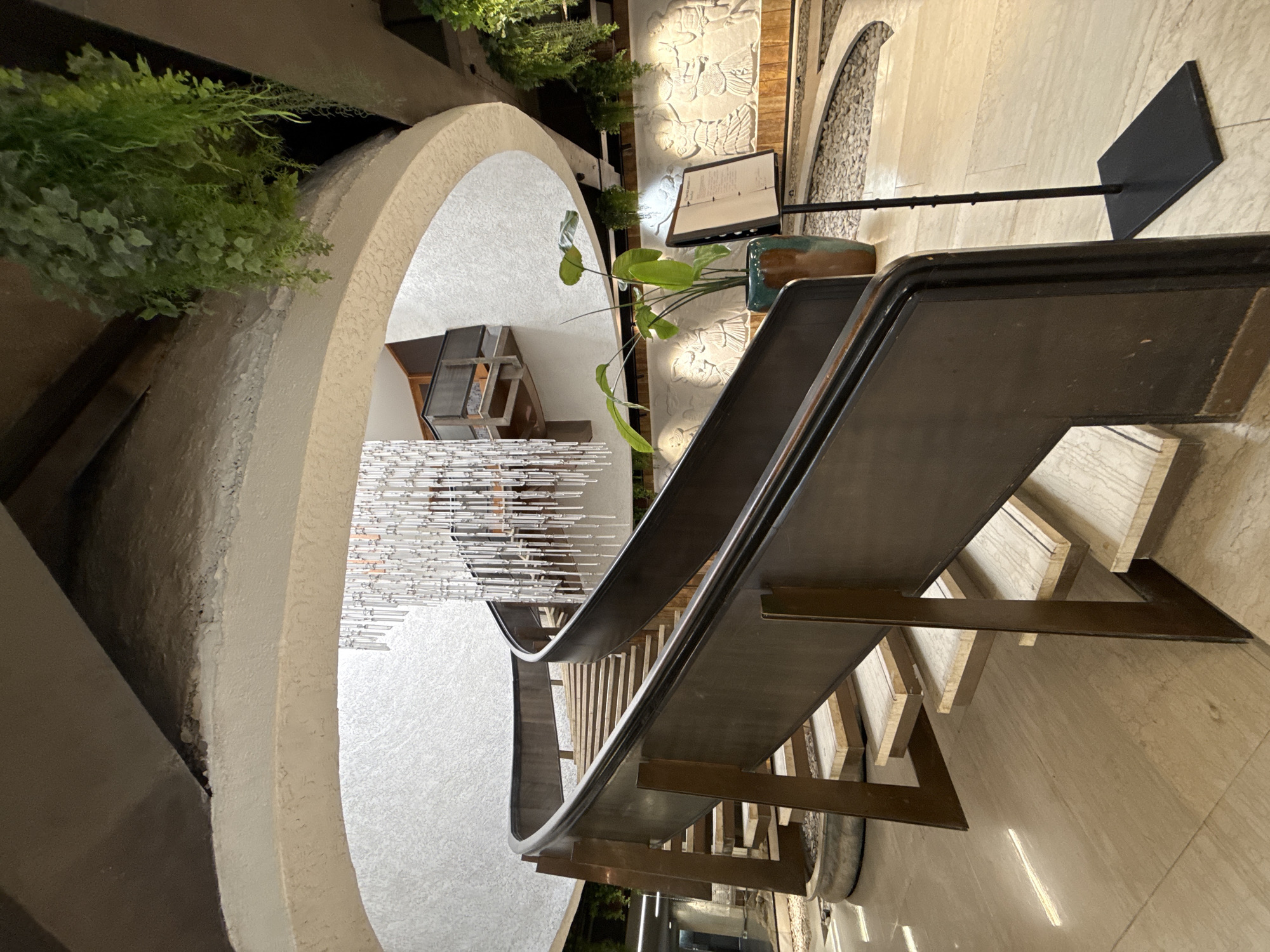 The image depicts an interior view of a building, featuring a grand spiral staircase with wooden steps and dark metal railings. The staircase curves upwards towards a circular opening in the ceiling, from which a large, modern chandelier made of glass tubes hangs. Lush green plants are suspended from the ceiling and edges of the structure, adding a touch of nature to the scene. Below the staircase, a wall features intricate bas-relief carvings of figures in traditional attire. A potted plant stands to the right of the staircase, next to a standing menu holder. The floor is made of polished light-colored stone tiles. The overall ambiance suggests a hotel lobby or a similar public space within a resort or establishment. The location is identified as Yalta, Ukraine. There are no people visible in the image.