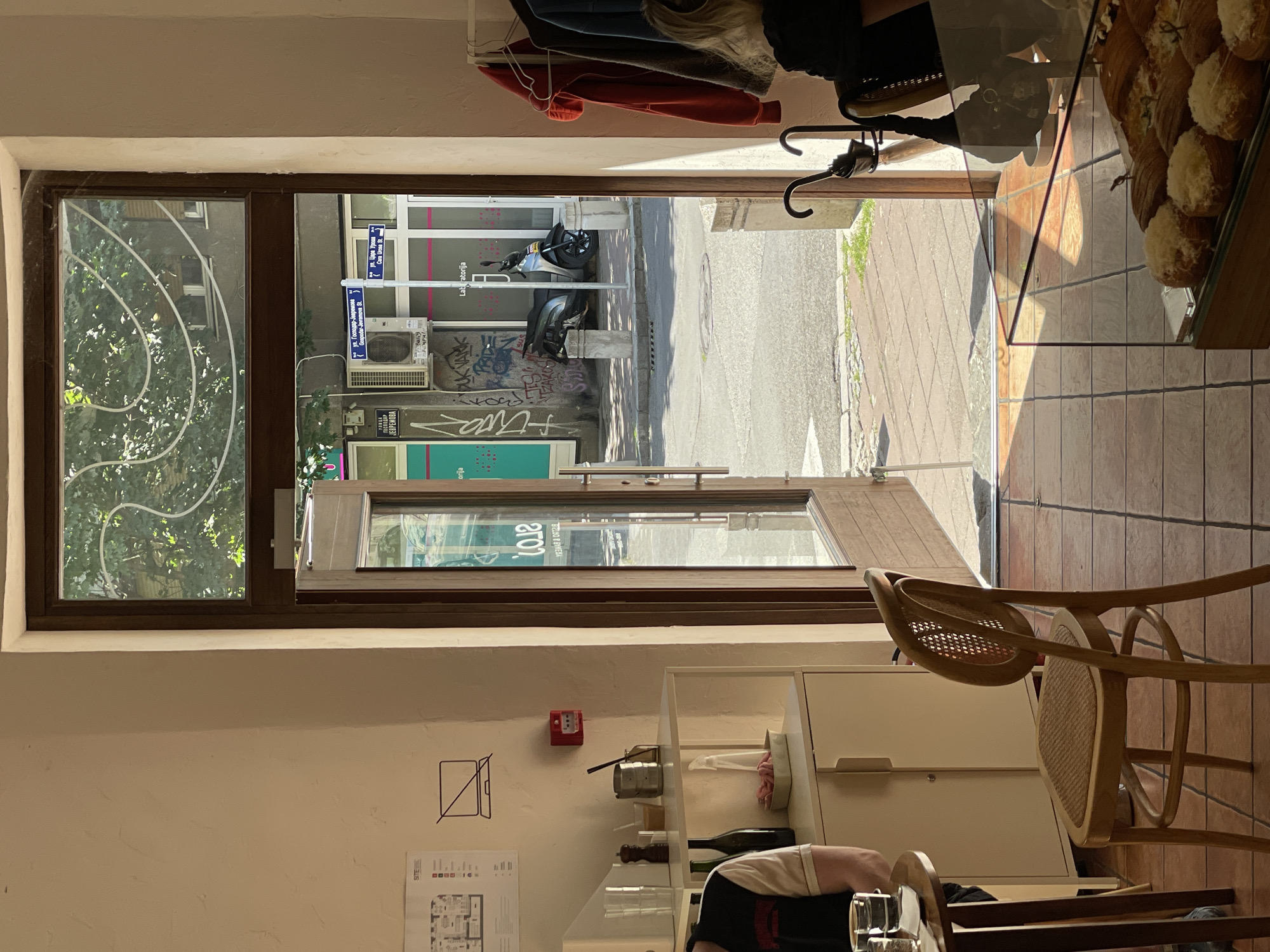 The image shows the interior of a shop or cafe in Belgrade, Serbia, looking out through an open door. Outside the door, a street scene is visible, with parked motorcycles, a building with graffiti, and street signs indicating "ул. Господар-Јевремова" and "ул. Цара Уроша" (Gospodar-Jevremova Street and Cara Urosa Street). The shop's interior features baked goods in a display case, a wicker chair, and storage cabinets. Clothes are hanging near the doorway. A person's shoulder and arm are visible, suggesting they are seated at a table within the cafe.