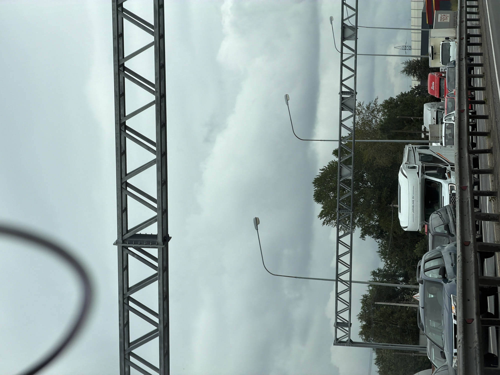 The image depicts a traffic jam on a cloudy day. Several cars and trucks are visible, including a white RV with "JIANGSLING RV" branding, a red truck, and several other vehicles. The scene appears to be on a highway or a major road, indicated by the overhead gantry structures and streetlights. The overall environment is overcast, suggesting a typical St. Petersburg weather condition. The presence of traffic and road infrastructure suggests an urban or suburban setting within Saint Petersburg, Russia.