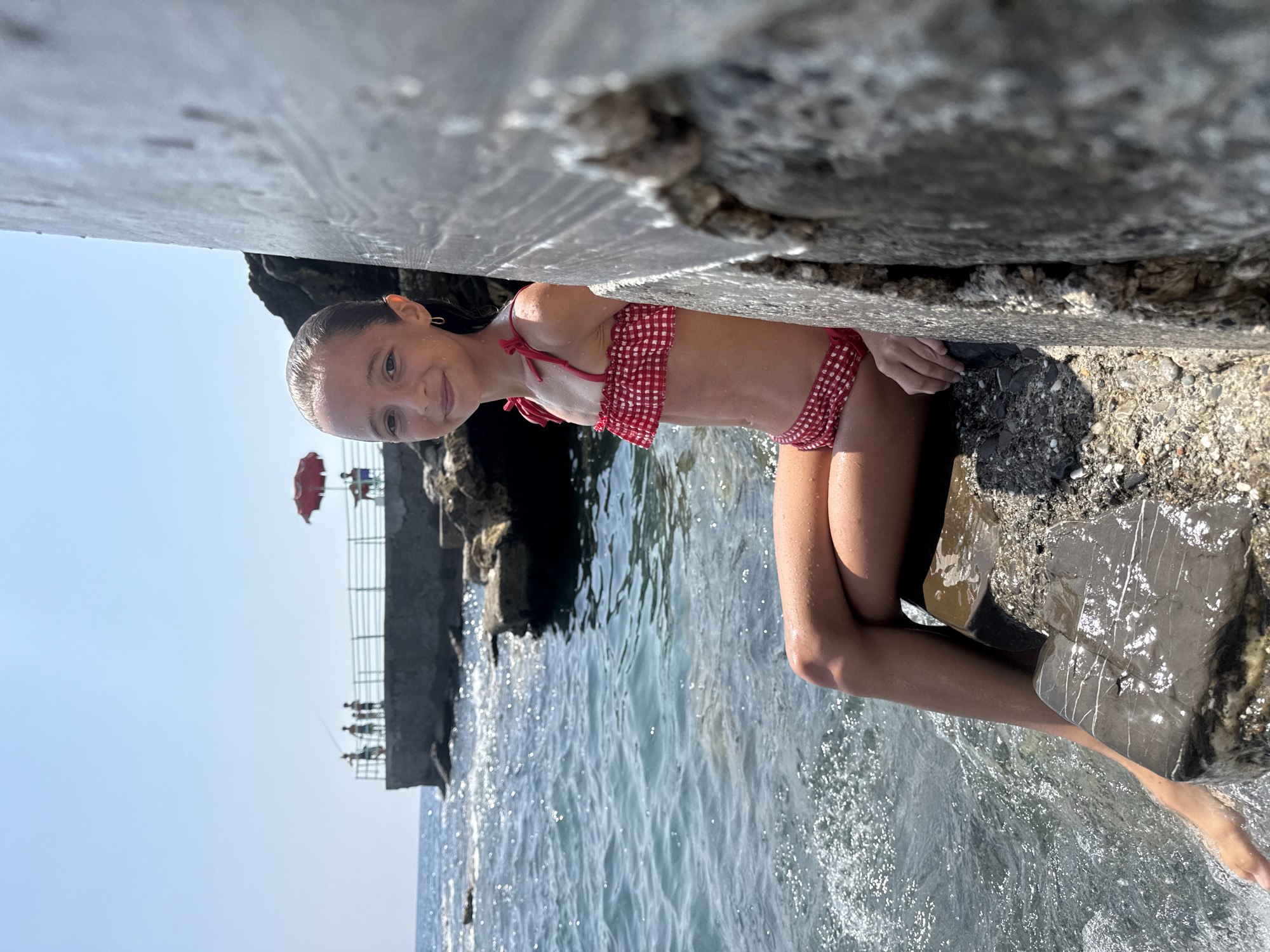 The image shows a young girl in a red and white checkered bikini sitting on a rocky shore with the sea to her left. She is looking at the camera with a slight smile. Her hair is wet, and she appears to be enjoying the day. The water is clear and blue, with small waves lapping at the shore. In the background, there is a pier with a red umbrella and a few people. The sky is clear and blue, suggesting it is a sunny day. The overall scene is one of a pleasant summer day at the beach. The image is oriented vertically.