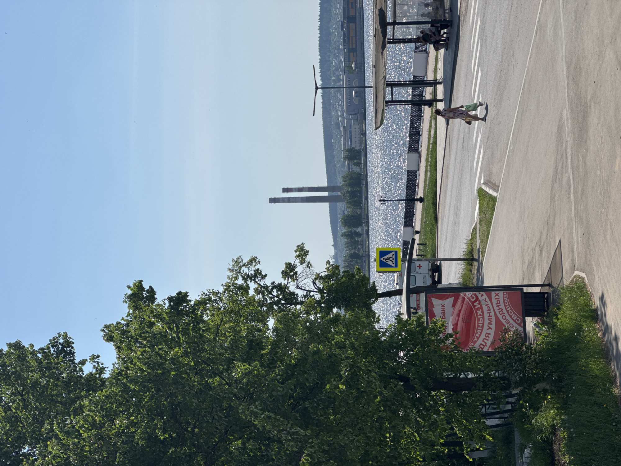 The image depicts a street scene in Votkinsk, Russia, adjacent to a body of water, likely the Votkinsk Reservoir. Two industrial chimneys are visible on the opposite shore. A pedestrian is crossing a crosswalk. A pedestrian crossing sign is present. There is an advertisement sign in Russian. The foreground is dominated by green trees. The sky is clear and blue. Along the waterfront, there is a bus shelter.