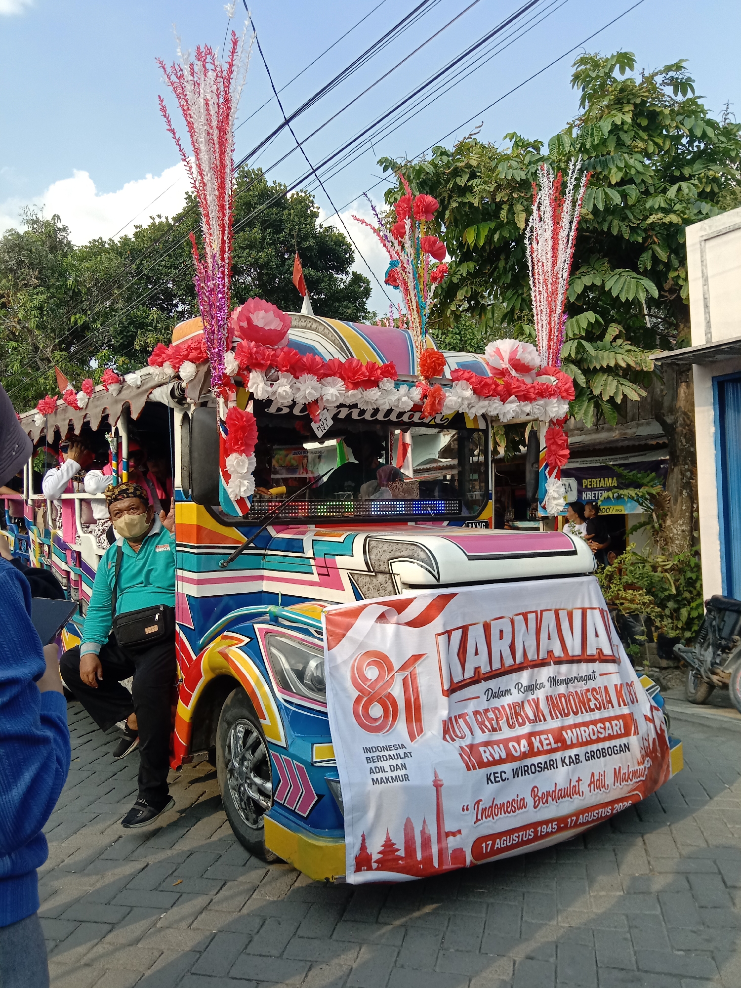 The image shows a colorful, decorated vehicle, likely a "odong-odong" or similar open-air transport, parked on a brick-paved street. The vehicle is adorned with red and white floral decorations, streamers, and a large banner. The banner prominently displays "KARNAVA" and text in Indonesian, indicating it is for a celebration in Wirosari, Indonesia. Text on the banner reads: "Dalam Rangka Memperingati HUT REPUBLIK INDONESIA KE 81 RW 04 KEL. WIROSARI KEC. WIROSARI KAB. GROBOGAN 'Indonesia Berdaulat, Adil, Makmur' 17 AGUSTUS 1945 - 17 AGUSTUS 2026". A man wearing a light blue shirt and a beige mask is seated at the front of the vehicle, looking towards the camera and smiling. Other people are visible inside the vehicle, some obscured. Green trees and power lines are in the background, suggesting an outdoor, possibly residential or community street setting. The overall atmosphere appears festive, with the decorated vehicle and the banner suggesting a public event or parade.