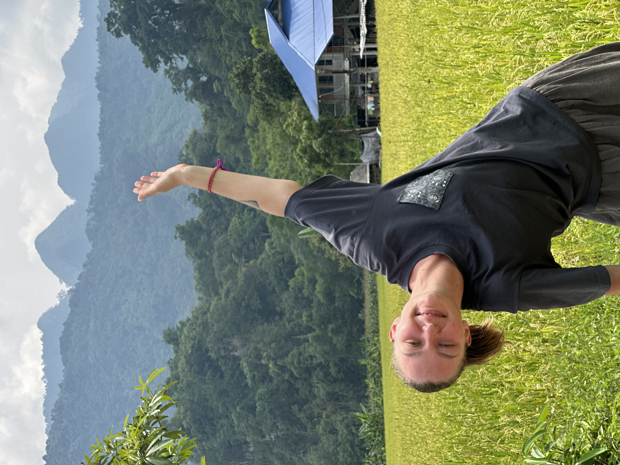 A vibrant, rotated full-body photograph captures a smiling individual amidst a picturesque rural landscape in Ngọc Đường Commune, Vietnam. The subject, appearing to be a young woman or adult, is positioned horizontally across the frame, looking towards the viewer with a broad, cheerful smile, revealing visible braces. She wears a dark grey t-shirt featuring a distinctive patterned patch on the left side, along with dark grey pants or a skirt. A pink braided bracelet adorns her right wrist, and her right arm is extended outwards with an open palm towards the distant scenery.
The background is rich with natural elements: a vast, bright green field of crops, likely rice paddies, stretches across the right side of the frame. Beyond the crops, lush, forested mountains rise majestically under a partly cloudy sky, creating a layered, serene backdrop. Nestled among the trees in the background, a multi-story building with a striking bright blue metal roof is partially visible.
The scene is illuminated by natural daylight, suggesting a pleasant time of day, possibly late morning or early afternoon. The individual's happy expression conveys a sense of contentment and engagement with the beautiful surroundings. The unique angle of the shot adds a dynamic and inviting quality to the image. There is no visible text in the photograph.