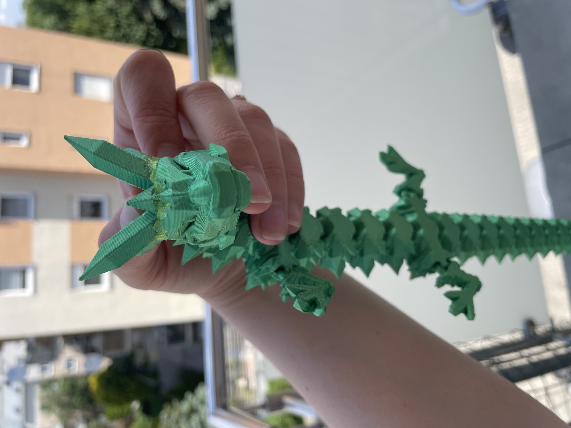 A close-up image depicts a person's right hand holding a green, articulated 3D-printed flexible dragon toy. The hand exhibits light skin with short, unpolished fingernails, holding the dragon by its upper body, with the dragon's head facing the viewer. The dragon is a solid, opaque green with distinct FDM print layer lines visible throughout its segmented body, faceted head, and pointed dorsal spikes/horns. Slight filament stringing or residue is present around the base of the head's spiked features. The background is a blurred outdoor scene under bright daylight. Prominent in the mid-ground is a multi-story residential building, primarily light beige with an orange-colored section, featuring several rectangular windows, some with white frames and closed blinds. Additional residential structures and green deciduous trees and bushes are visible further in the distance. A metallic railing, indicative of a balcony or terrace, is partially visible in the lower portions of the frame. The overall environment suggests an urban or suburban residential setting.