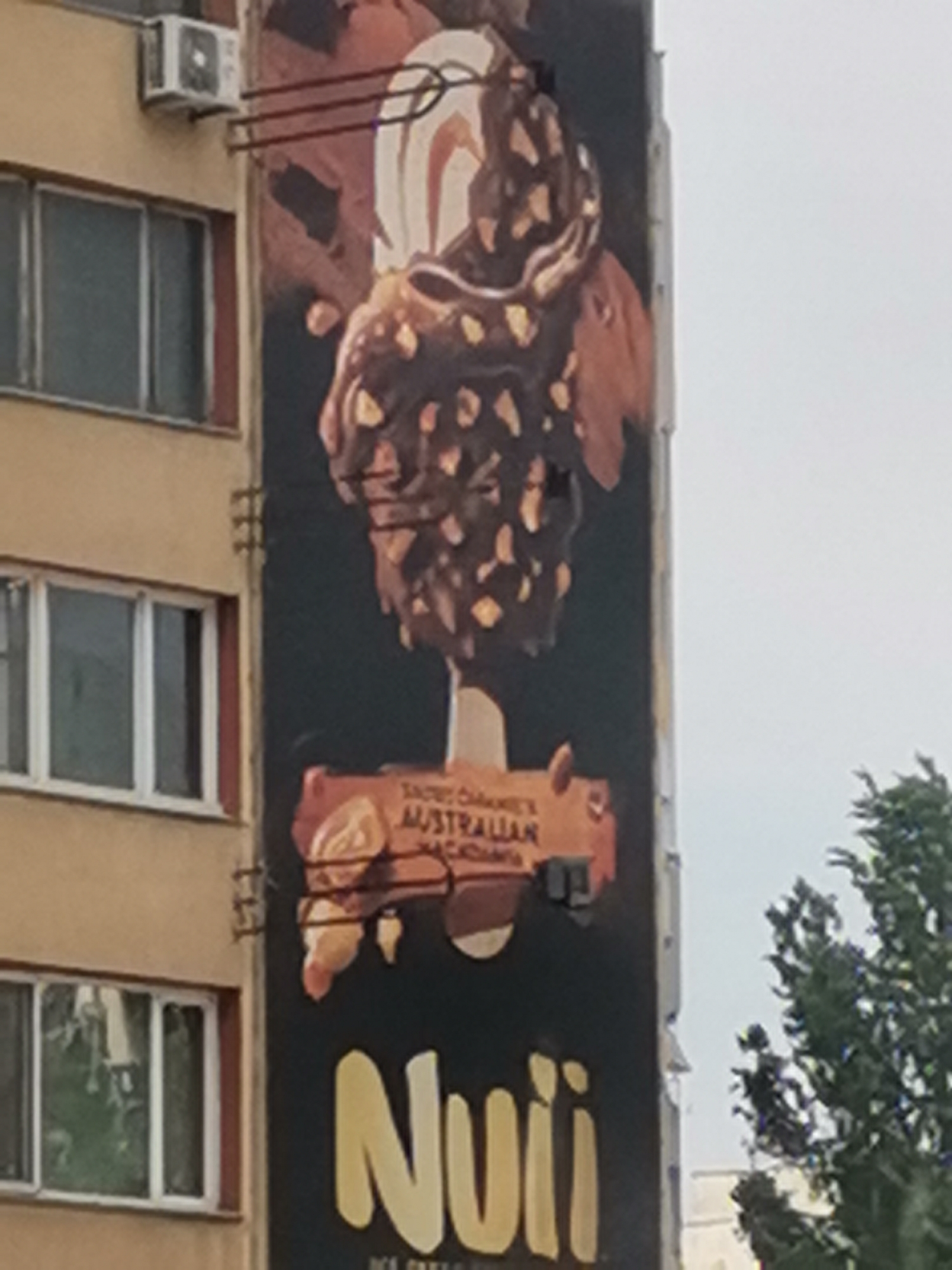 The image shows a large billboard advertisement for "Nuui" ice cream, featuring a prominent depiction of a chocolate-covered ice cream bar with nuts. The billboard is mounted on the side of a building, which appears to be a residential apartment building, with visible windows and an air conditioning unit. A section of a green tree is visible on the right side of the frame. The advertisement includes text that reads "SAINT GEORGE'S AUSTRALIAN MACADAMIA." The billboard is vertical and covers a significant portion of the building's facade. The overall scene suggests an urban environment, likely in a city like Sofia, Bulgaria, given the visual context of the building architecture and typical urban advertising.