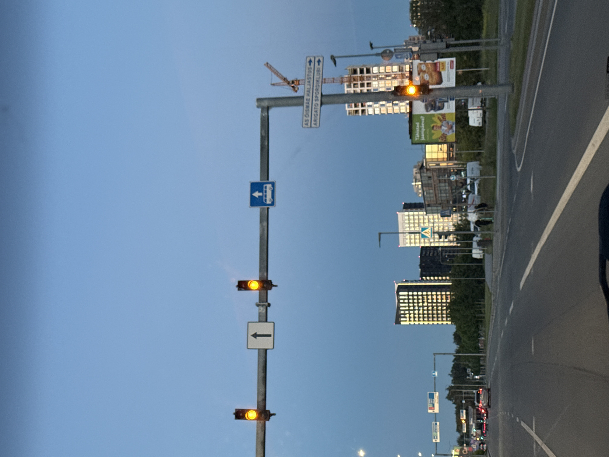 The image shows a road in Tallinn, Estonia, at dusk. The sky is a clear, pale blue. In the foreground, there is a two-lane road with white lane markings. In the background, there are several modern buildings, some of which are illuminated. Traffic lights are visible, and one of them is illuminated amber. A construction crane is also visible near one of the buildings. Several cars are driving on the road. Two signs are visible on a pole, one pointing to "AS Oismae Haljastus" and the other to "Arigato Spordiklubi". There are also some billboards visible in the background.