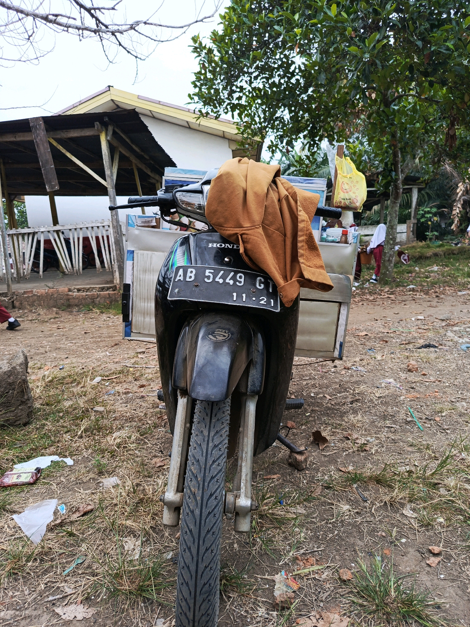 In Megang Sakti II, Indonesia, a parked motorcycle with a sidecar is the central subject. The motorcycle is black, with the license plate "AB 5449 CI 11-21" visible. A brown jacket is draped over the handlebars. The motorcycle is parked on a dirt and grass surface littered with debris, including plastic bags, wrappers, and leaves. In the background, a simple structure with a wooden roof and white walls is partially visible, along with trees. Several people, including two wearing white and red clothing, are present in the background, one of whom is standing near a tree and another is partially visible near the structure. The overall scene depicts an outdoor environment that appears to be somewhat unkempt.