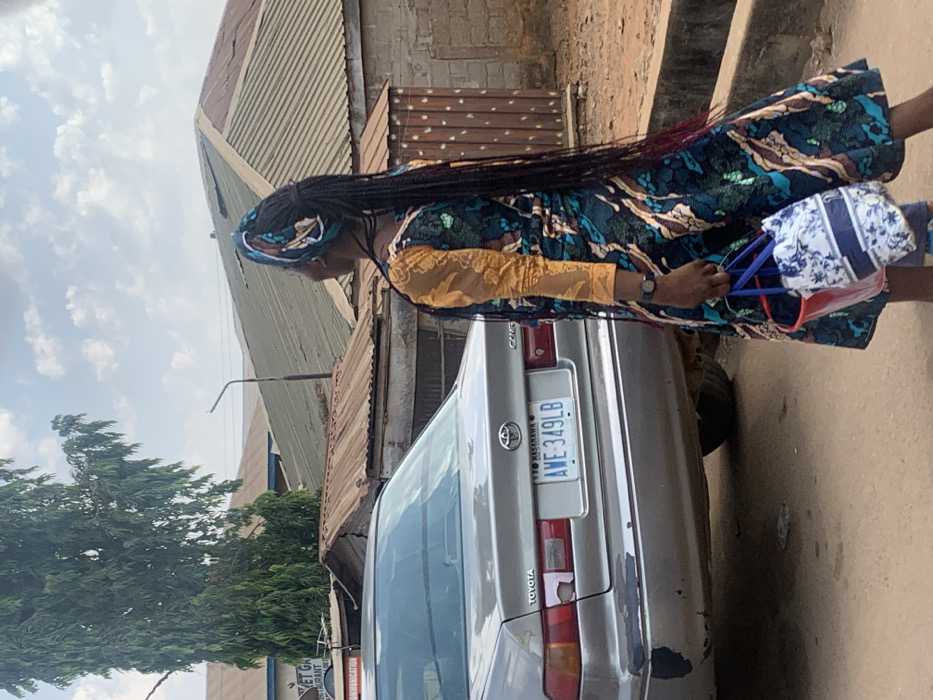 The scene depicts a street in Egbedi, Nasarawa State, Nigeria, under a partly cloudy daytime sky. In the foreground, a person, viewed from behind and slightly to the left, walks or stands on an unpaved, reddish-brown dirt surface. The individual has dark skin, wears a long, patterned dress featuring a teal, brown, and gold design with a golden lace long sleeve visible on the right arm, and a matching or complementary headwrap. Long, dark braided hair extends down the back, with some strands showing reddish tips. A dark-colored wristwatch is present on the left wrist. The individual carries two bags in the left hand: a white bag with a blue floral or abstract pattern and a smaller, translucent red bucket-style bag.
To the left of the person is the rear-left section of a silver/light gray Toyota Camry sedan. The car displays a Nigerian license plate: "NASARAWA AWE-349LB". The rear bumper shows significant peeling paint, revealing dark underlying material, and the left taillight area has a faded or damaged pinkish patch.
In the mid-ground and background, several buildings with corrugated metal roofs, mostly brown and rusty, are visible. One building on the left has a partially obscured sign that reads "VET GA..." or "VET GK..." and "COMMUNICATION". A tall tree with dense green foliage stands to the far left, partially obscuring the upper portion of a building. A utility pole or street light standard with an upward-curving arm is visible between the buildings and the person.