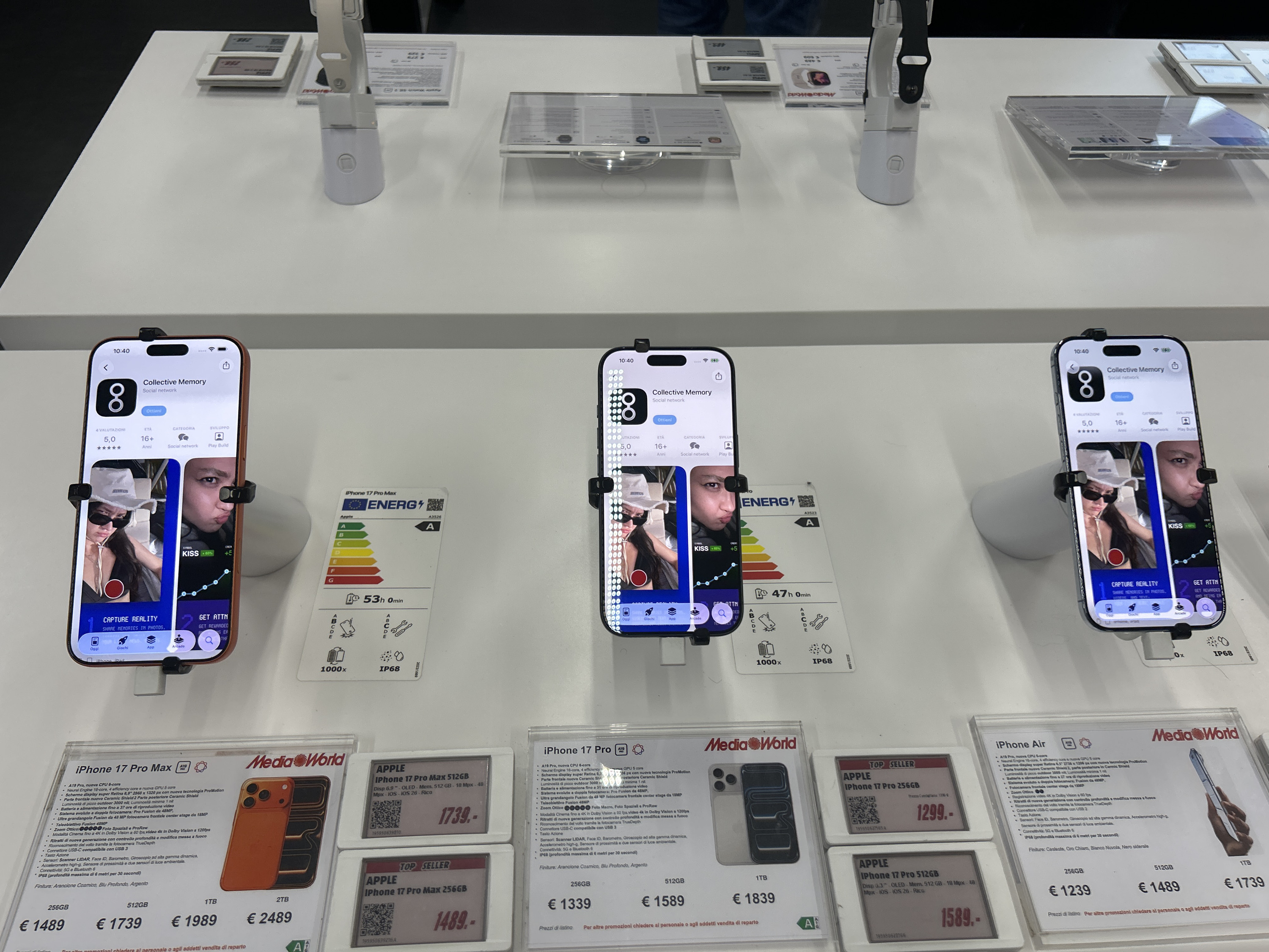 The image depicts a well-lit, white retail display counter within a MediaWorld store located in Lipomo, Italy. Three smartphones, identified as iPhones (specifically, iPhone 17 Pro Max, iPhone 17 Pro, and iPhone Air models based on adjacent price cards), are showcased vertically, secured by black, two-pronged security mounts.
Each phone's screen displays the identical interface of an app store page for "Collective Memory," a social network application with a 5.0-star rating and a 16+ age recommendation. The app's interface shows two distinct image previews: one featuring a person in a white bucket hat with "JACQUEMUS" text and sunglasses, and another of a person making a pouting or "duck face" expression.
Beneath the phones, the white counter holds various clear acrylic product information cards and digital price tags. These cards detail specifications, storage capacities (256GB, 512GB, 1TB, 2TB), and prices in Euros for the displayed iPhone models, with prices ranging from €1239 to €2489. Several cards also feature "Energia" labels indicating energy efficiency (Class A) and estimated battery life (e.g., 53h 0min, 47h 0min), alongside symbols for features like 1000x zoom and IP68 water resistance. Digital tags prominently display "APPLE" and "TOP SELLER" alongside prices.
In the upper background, additional white security stands and information cards are visible, suggesting other products such as smartwatches are also on display. The overall environment is a clean, organized electronics retail setting.
