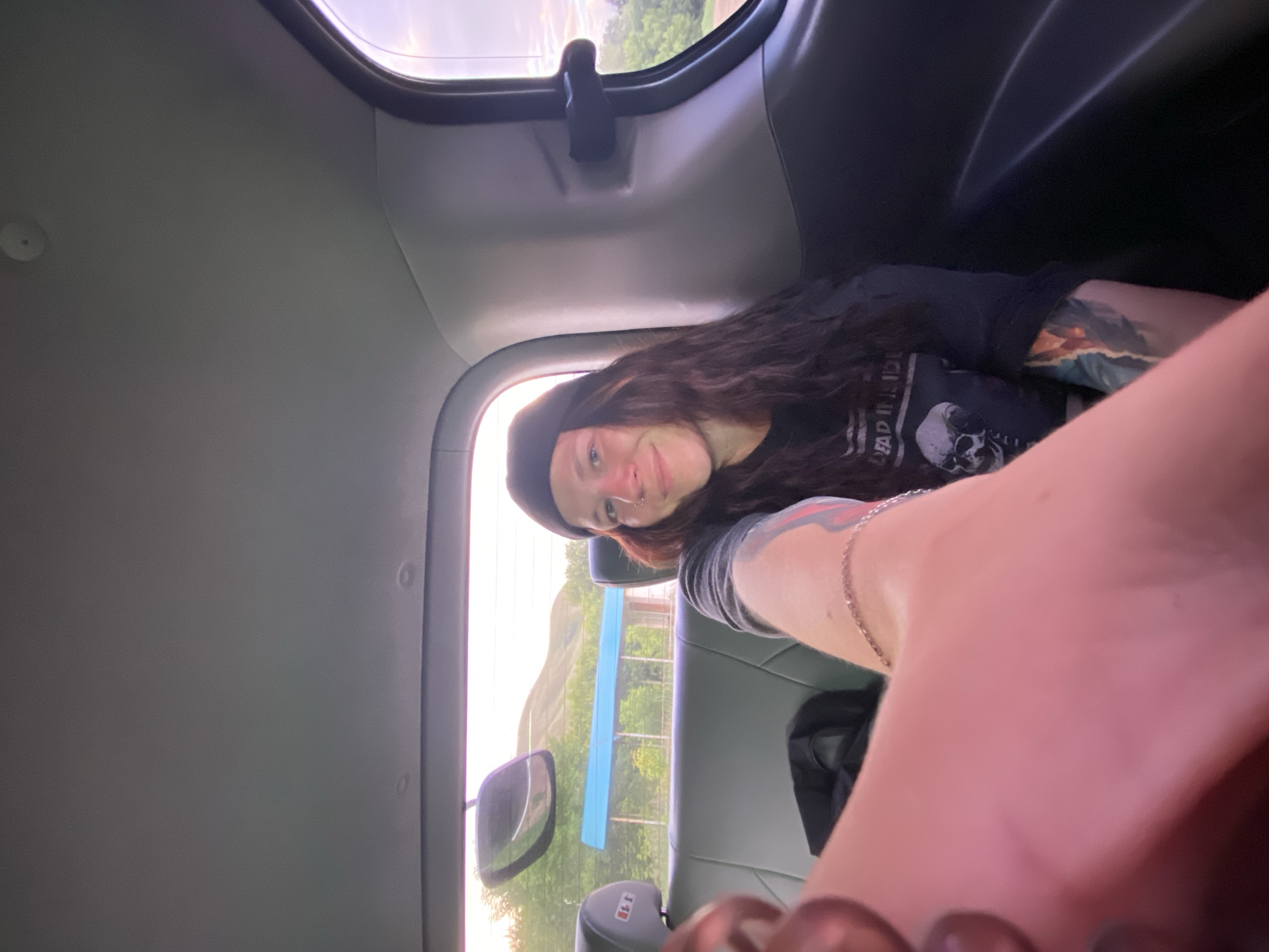 A young person with long, dark, wavy hair and a black beanie is sitting in the backseat of a vehicle, taking a selfie. They are wearing a black t-shirt with a skull graphic and the words "DEAD INSIDE" visible. The person has tattoos on their arms. The interior of the vehicle is visible, including the gray leather seats, a rearview mirror reflecting a hilly landscape with a blue-roofed structure, and the vehicle's ceiling and side panels. The background outside the windows shows a slightly hazy, overcast sky and green foliage. The location is the city of Verkhneozernoye, Russia.