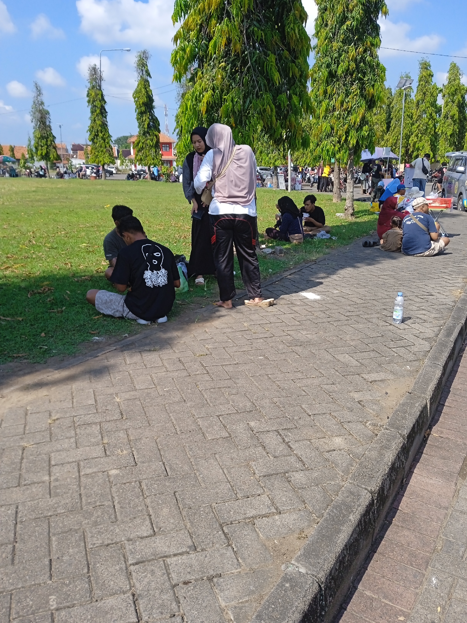 People are sitting on grass beside a paved area with water bottle. The background shows trees, vehicles, and buildings. The location is in Kebondalem, Indonesia.