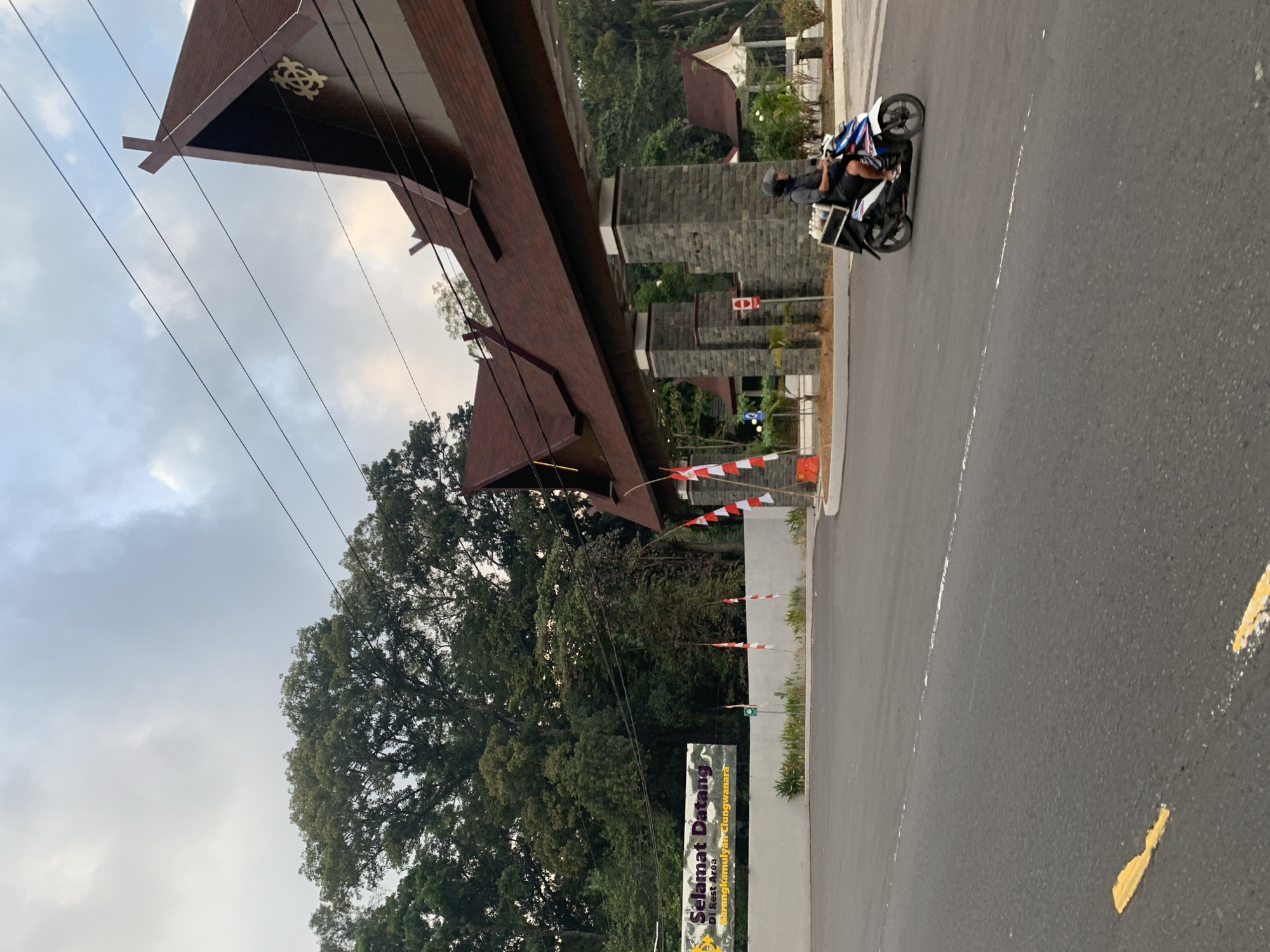 The image captures a view of an entrance to a rest area in Karangkamulyan, Indonesia. A large, ornate wooden archway with a traditional architectural style marks the entrance. The archway is supported by stone pillars. In front of the archway, there is a white wall with a banner that reads "Selamat Datang Di Rest Area Karangkamulyan Clungwanara" in Indonesian, meaning "Welcome to Karangkamulyan Clungwanara Rest Area." Several Indonesian flags, red and white, are mounted on poles along the wall.
A man is riding a white and blue scooter on the road, moving away from the camera. He is wearing a helmet and has a black cargo box attached to the back of the scooter, filled with white items. The road in the foreground is asphalt and has yellow and white lane markings. In the background, there are lush green trees and some buildings with similar traditional roof designs as the archway. The sky is overcast with clouds. The overall scene depicts a welcoming entrance to a rest stop, with a person on a scooter as the only visible human subject in motion.
