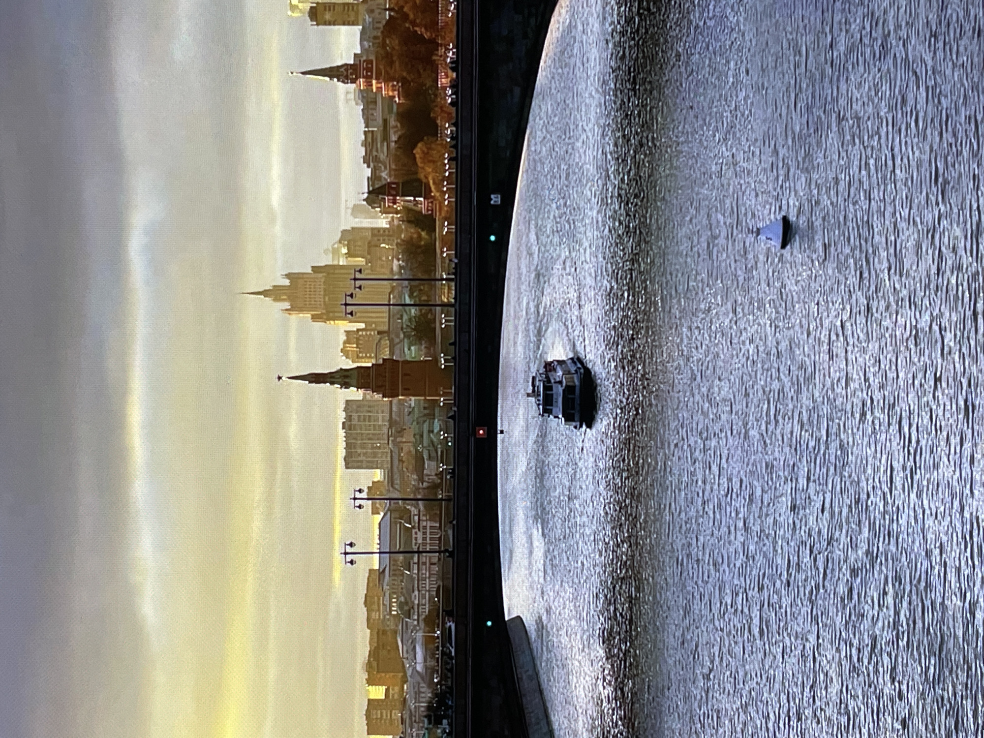 The image depicts a cityscape with a river in the foreground. A medium-sized white boat with a dark roof is sailing on the river. Further downriver, a white and black buoy is visible. A bridge spans the river, with lampposts lining its top. The background features a city skyline with several tall buildings, including a prominent skyscraper and a spire with a star on top. The sky is overcast with a warm, diffused light, suggesting either sunrise or sunset. The environment appears to be urban. The location is identified as Pushkino, Russia.