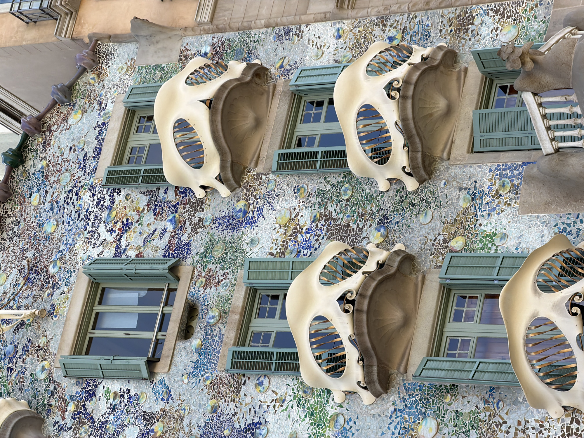 The image shows a close-up, upward-angle view of the facade of Casa Batlló, a building located in Barcelona, Spain, on Passeig de Gràcia. The facade is covered in a mosaic of colorful broken ceramic tiles, predominantly blues, greens, whites, and yellows, with some circular iridescent elements that resemble bubbles. There are several windows with green wooden shutters, some of which are open. Each window has a unique, bone-like balcony with a swirling, organic shape and brass-colored railings that are visible through the openings. The balconies appear to be made of a light-colored, molded material. The architectural style is characteristic of Antoni Gaudí's work, known for its curvilinear forms and use of vibrant colors and textures. The image is taken from a low vantage point, emphasizing the intricate details of the building's exterior. There are no people visible in the frame.