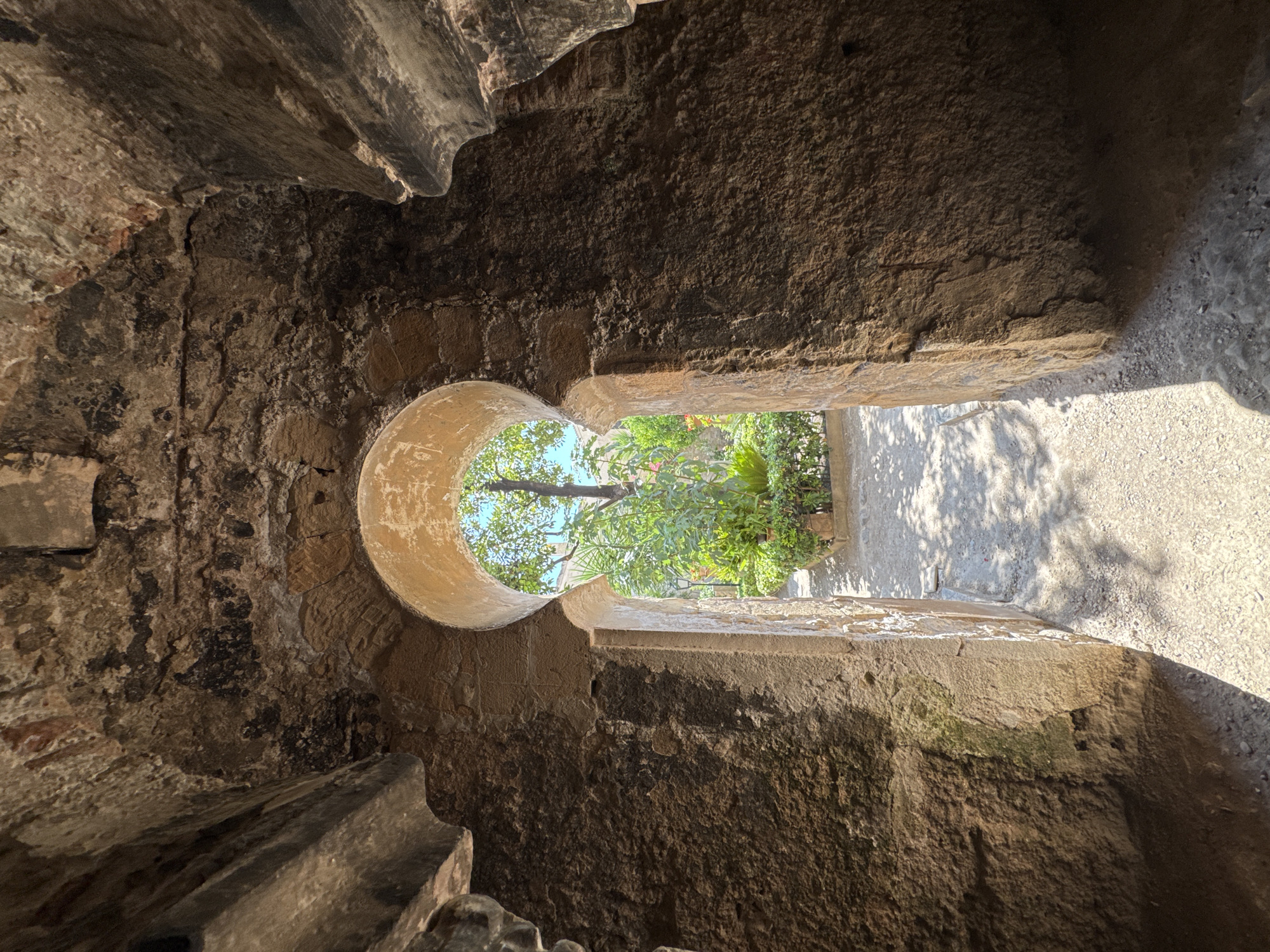 The image shows a view looking out from an arched stone doorway. The stone construction is rough and appears ancient, with visible textures and imperfections. The archway frames a sunlit courtyard or garden with lush green foliage and a large tree. Sunlight casts dappled shadows on the gravel path leading into the courtyard. The architectural style of the arch suggests a historical building, possibly a ruin or a well-preserved ancient structure. The setting appears to be a warm, Mediterranean climate. This view is from the Bellver Castle in Palma de Mallorca, Spain, specifically from one of its interior passages looking out towards the central courtyard.
