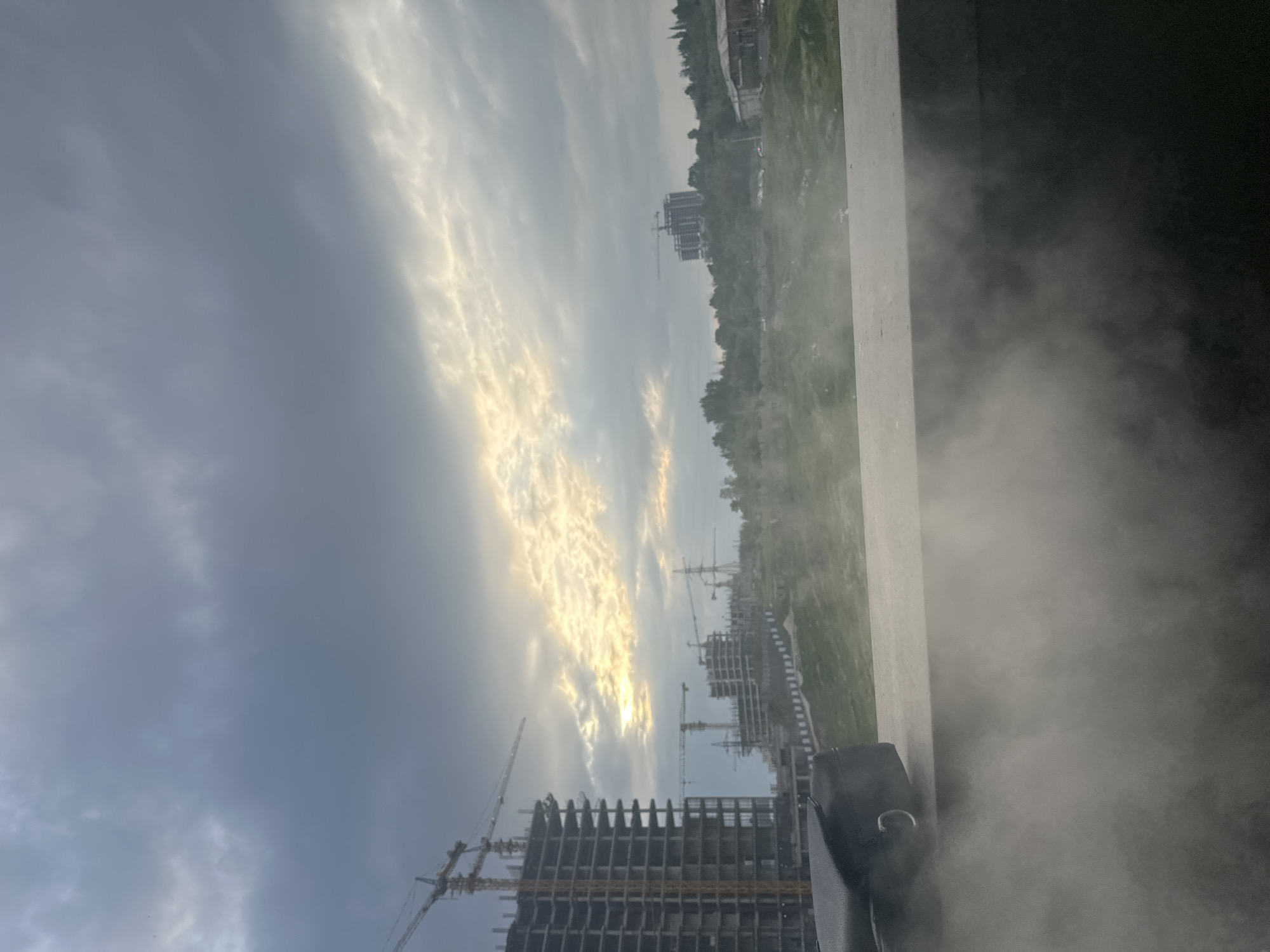 The image shows a construction site in a city. There are several buildings under construction, with cranes visible. The sky is cloudy with patches of sunlight breaking through. The location is Unknown, Unknown. The foreground is blurry, obscuring some details.