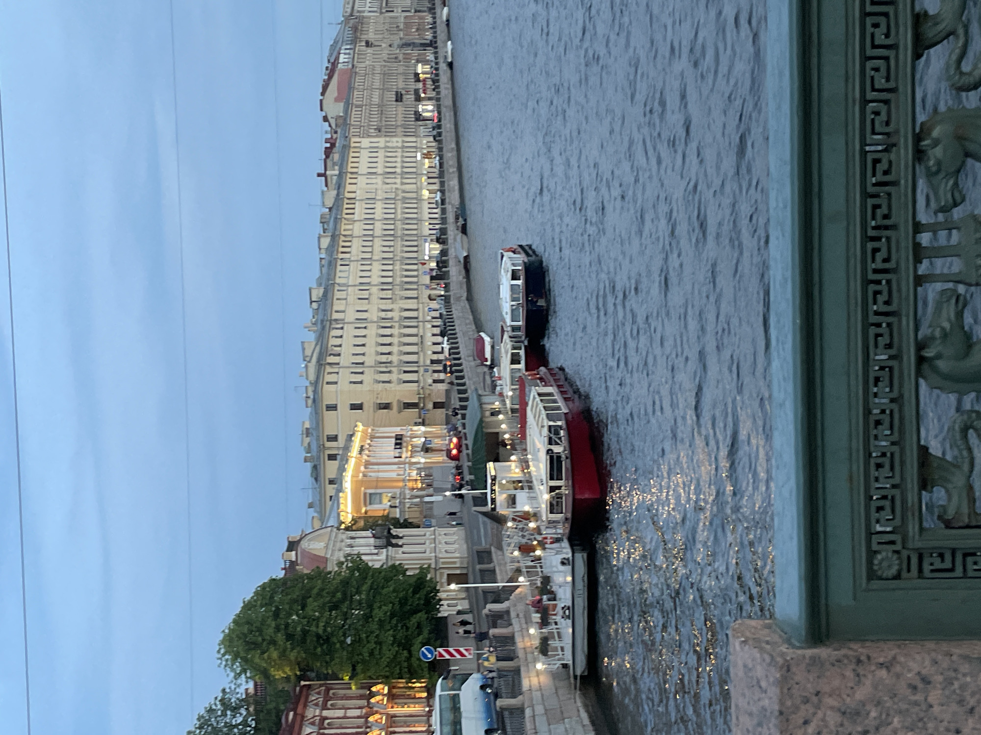 In Saint Petersburg, Russia, at twilight, a wide river flows with gentle ripples reflecting the dim sky. Along the embankment, a row of historic buildings with cream-colored facades stretches into the distance. Many windows glow with warm light, suggesting interior illumination. Several tour boats are docked along the river's edge. One is a red and white vessel, its deck illuminated by strings of lights. Adjacent to it, a smaller white boat is also lit. Further down, a dark-hulled boat is tied up. On the embankment, a few trees provide a touch of green against the architecture. A white bus is parked near the left side of the image, and several people are visible walking along the sidewalk. In the foreground, a section of a bridge railing is visible, made of dark green metal with intricate scrollwork and horse-shaped decorative elements. The overall atmosphere is tranquil, capturing the early evening ambiance of the city.
