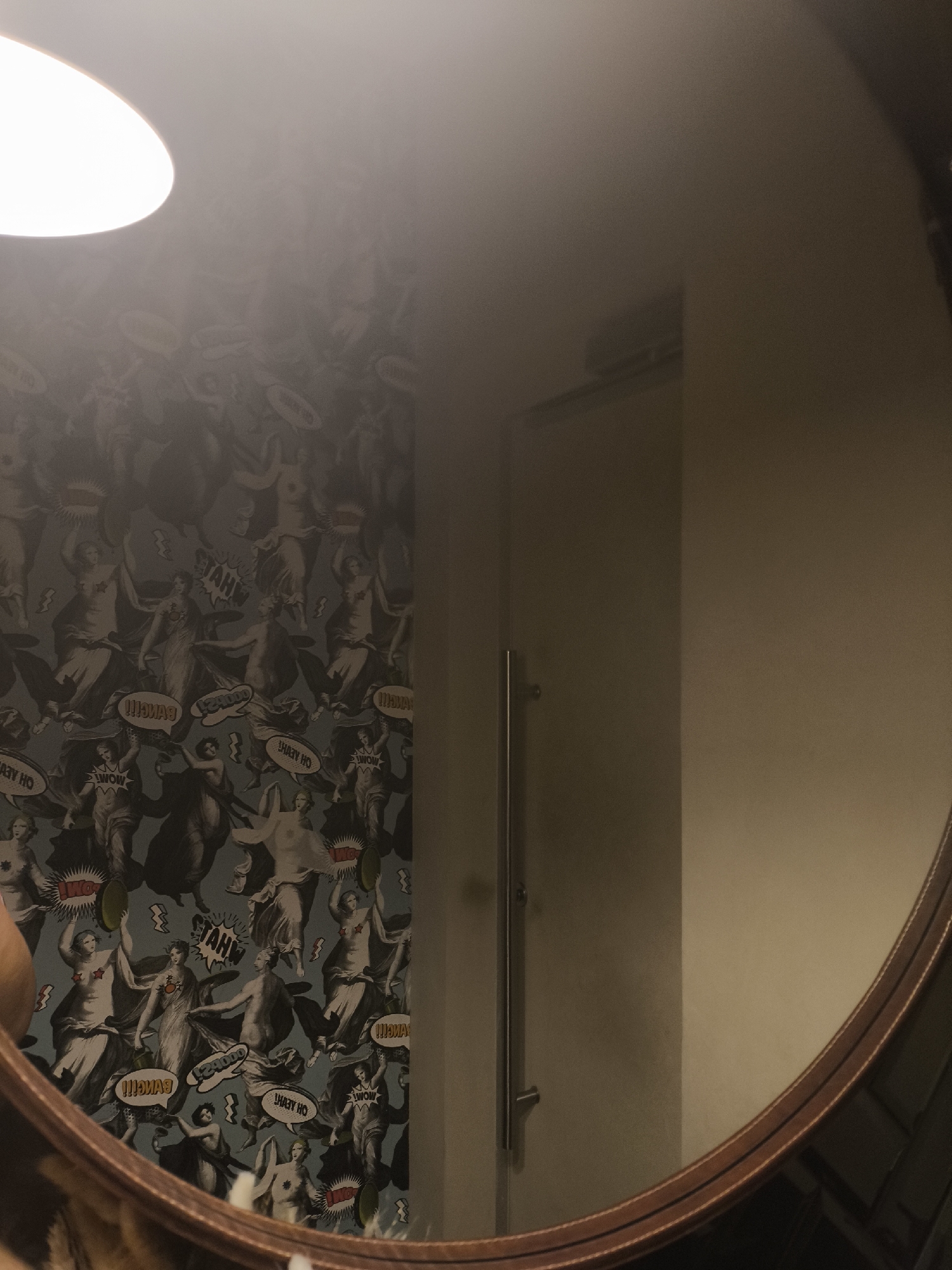 The image displays a circular mirror reflecting an indoor scene. The reflection prominently features a wall covered in elaborate wallpaper that combines classical art with pop art elements. The wallpaper pattern consists of numerous figures, reminiscent of ancient Greek or Roman goddesses, posed in various dynamic positions. Interspersed among these figures are speech bubbles containing words such as "WOW," "SYAH!", "BANG!!!", "OH YEAH HO," and "OMG," some of which appear reversed due to the mirror's reflection. Some figures have star-shaped or other symbolic coverings over their breasts. In the upper left portion of the reflection, a bright, circular overhead light fixture illuminates the area. To the right of the wallpapered wall, a plain, light-colored wall is visible, showing a vertical metallic fixture, possibly a handle or rail, reflected near its left edge. The mirror itself has a distinctive brown frame with decorative parallel lines or stitching. The bottom edge of the reflection shows an indistinct, blurry area, likely a partial view of the person holding the camera or their attire. There are no discernible people or explicit actions captured within the reflection, only the static environment. The scene lacks any identifiable markers or specific details that would allow for the determination of its precise location within the city of Izhevsk, Russia, or any other specific geographical area.