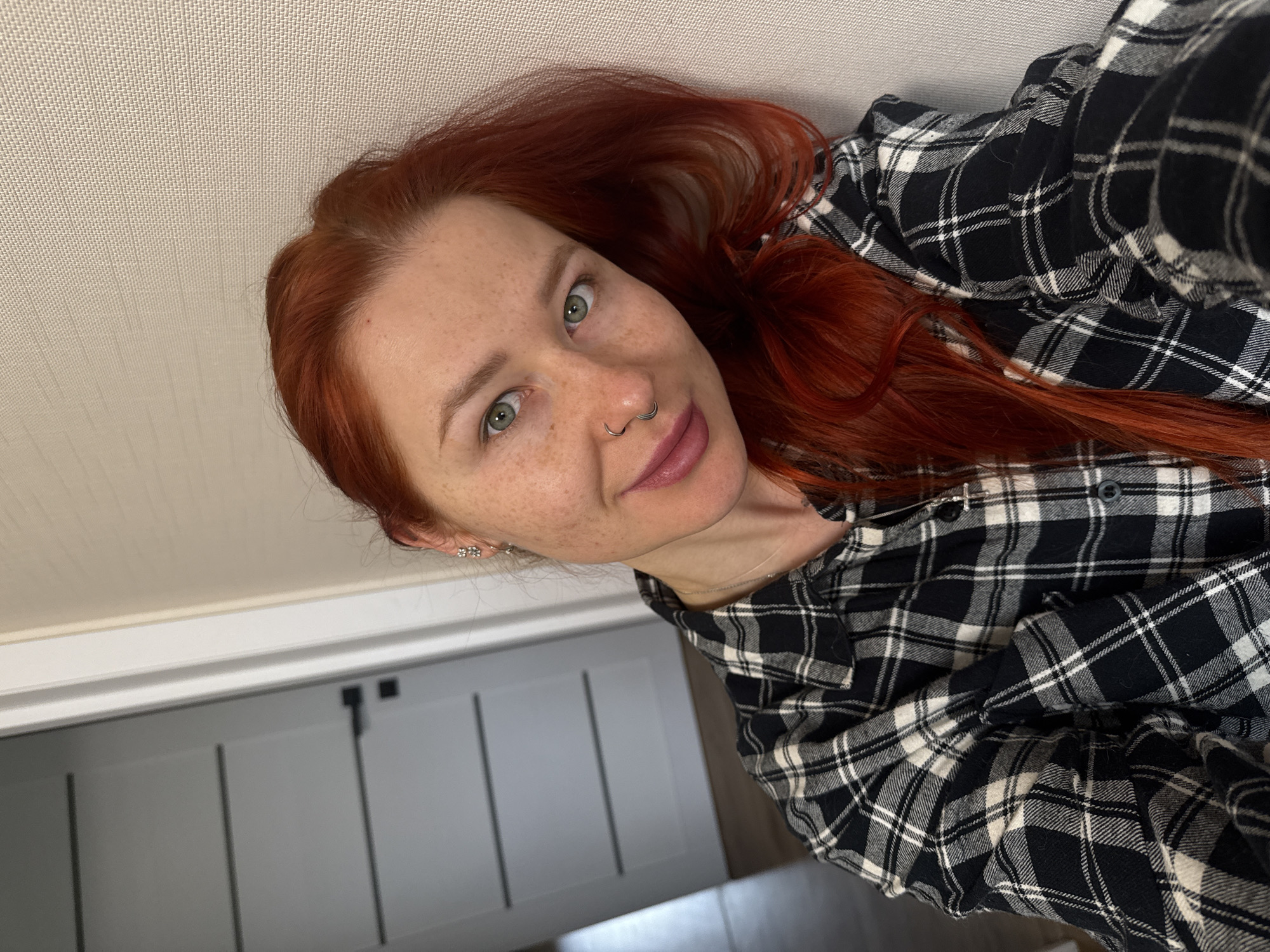 A selfie-style portrait features a young woman with vivid red hair and striking green eyes, looking directly at the camera. Her face is lightly freckled, and she has a septum piercing and small stud earrings in her left ear. Her lips are subtly made up in a muted pink or plum shade, and her expression is calm and direct.
She is wearing a black and white plaid flannel shirt, which appears soft and casual, with some of the fabric gathered around her. Her red hair cascades around her shoulders and upper back.
The background primarily consists of a light-colored, textured ceiling in the upper portion of the frame. Below that, part of a light gray wall or cabinet with vertical paneling details is visible, suggesting an indoor setting. The image is framed at an oblique angle, giving the impression that the subject might be reclining or the camera is tilted. The lighting is even and bright, consistent with indoor illumination. No text is visible in the image. The location context provided is Rostov-on-Don, Russia.