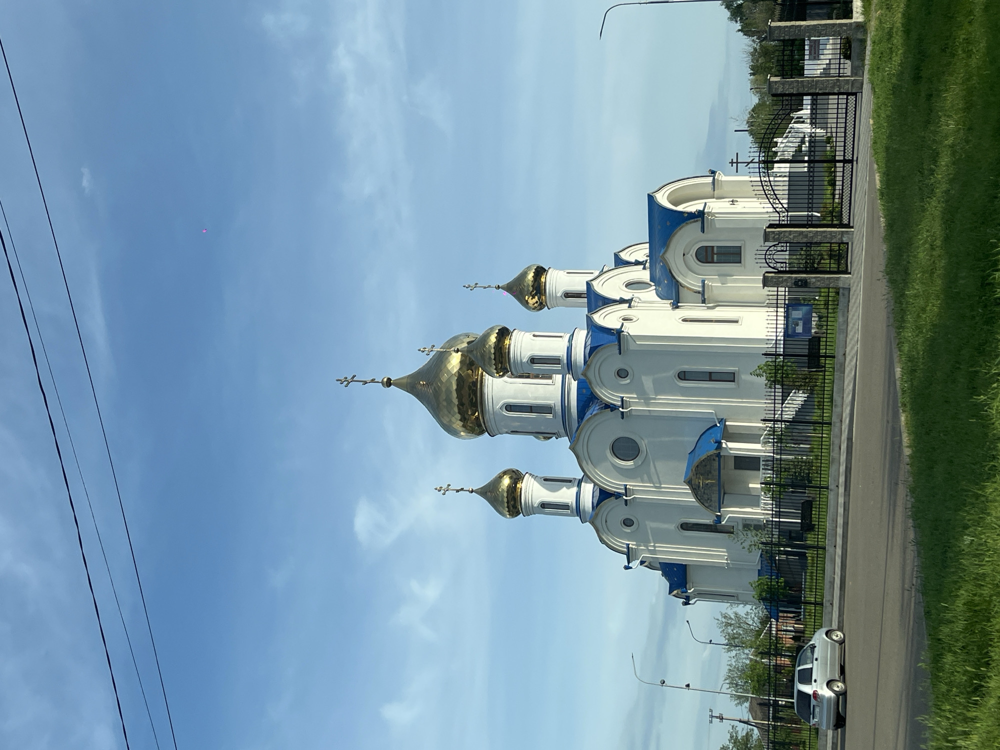 The image shows a white and blue Orthodox church with golden domes in Novopokrovskaya, Russia. A black wrought-iron fence borders the church. A road with green grass is on the right and a car parked on the road. Power lines are visible in the sky. A small pink spot is noticeable in the sky. The sky is blue with light clouds.