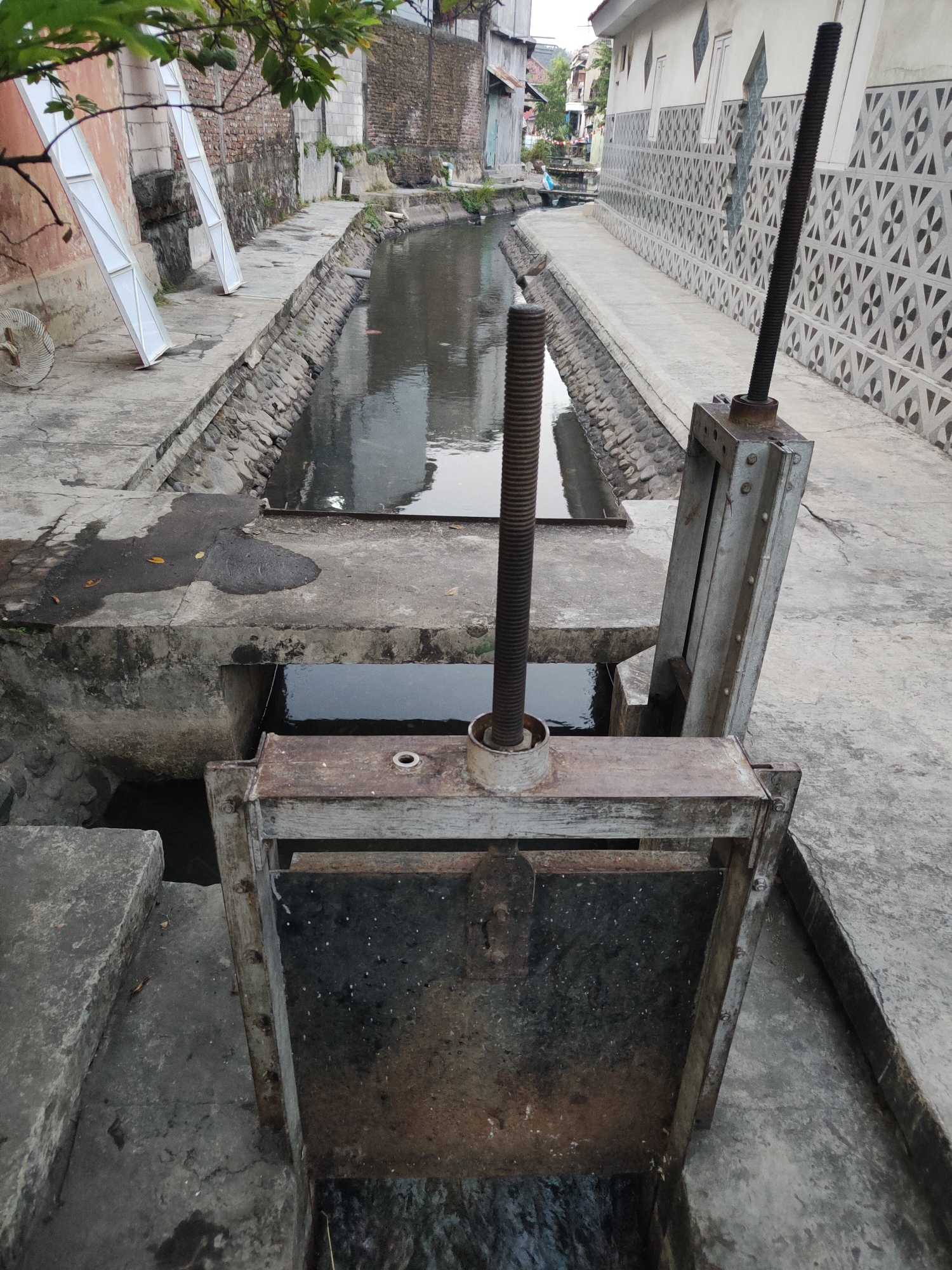 The image shows a canal or drainage ditch in Kauman, Indonesia. The canal is lined with concrete and stones. On the right side of the canal is a walkway with a decorative patterned wall. On the left side, there are buildings and some white panels leaning against a brick wall. In the foreground, a metal sluice gate mechanism is visible, with a threaded screw rod extending vertically. The gate itself is in a lowered position, allowing water to flow beneath it. The water in the canal appears murky. The scene depicts an urban environment with functional infrastructure for water management.