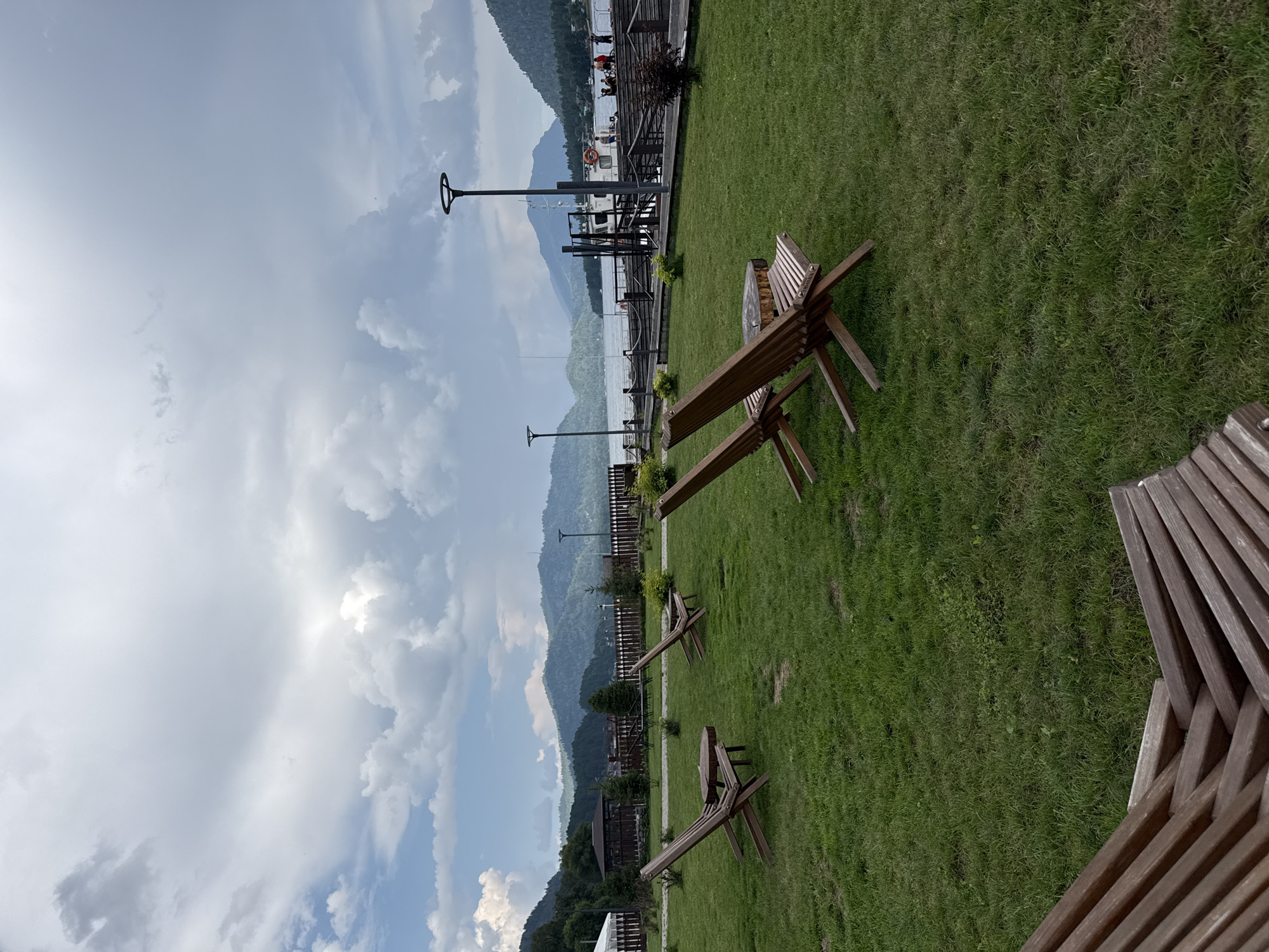 The image depicts a scenic outdoor area in Artybash, Russia, featuring a well-maintained grassy lawn dotted with wooden lounge chairs and a small wooden table. In the background, a wooden pier extends into a body of water, where a white boat with several people on board is docked. Beyond the water, a range of lush green mountains under a cloudy sky forms the backdrop. The overall atmosphere suggests a peaceful recreational setting, possibly a resort or a public park by the lake.
