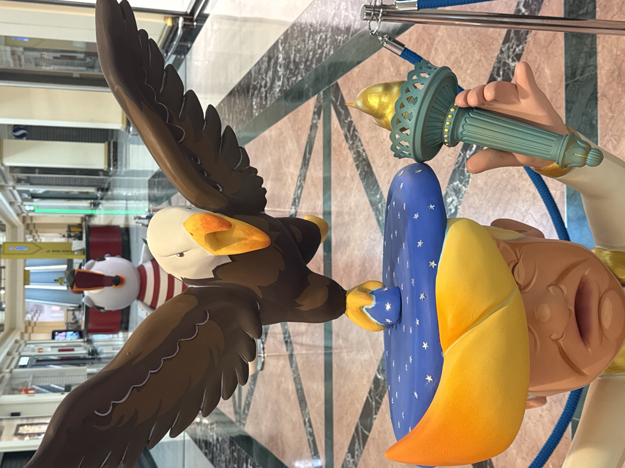 A low-angle medium shot captures a large, cartoonish sculpture of a bald eagle with outstretched wings, flying above a caricature of a man wearing a large, yellow, crescent-shaped hat adorned with white stars on a blue background. The man has a distressed expression, with furrowed brows and a gaping mouth. He is holding a teal-colored torch with a golden flame. The sculpture appears to be a public art installation, possibly in a shopping mall, given the tiled floor, storefronts, and railings visible in the background. The lighting is bright and even, typical of an indoor commercial space. The colors are vibrant and saturated, emphasizing the artificial nature of the sculpture.