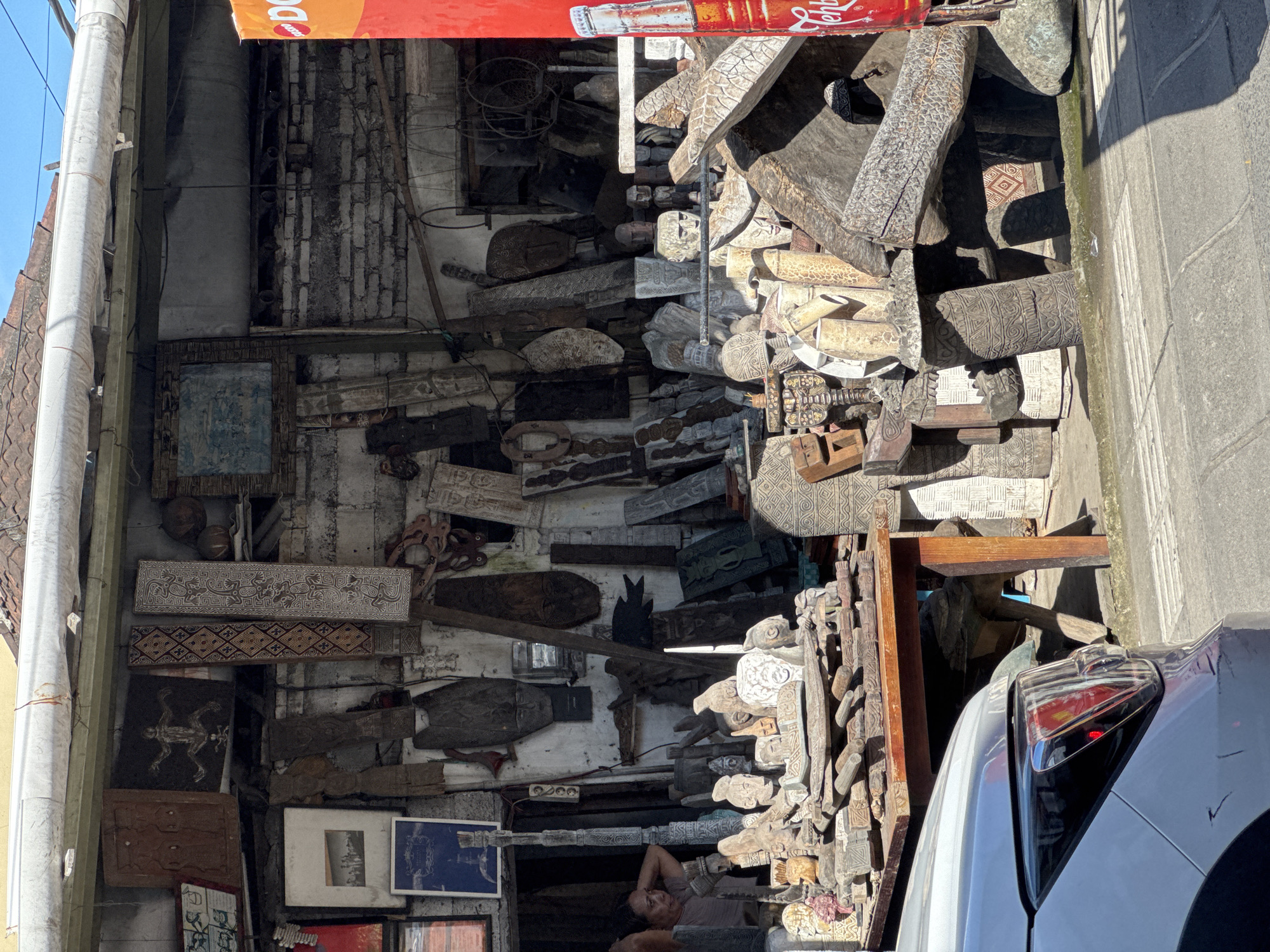 The image captures a densely packed outdoor or semi-outdoor space, likely a workshop or shop, filled with a multitude of carved wooden objects. The scene is illuminated by bright sunlight, casting sharp shadows.
Thousands of wooden carvings dominate the frame, ranging from long, flat planks adorned with intricate geometric patterns and stylized motifs to smaller, three-dimensional pieces resembling human faces, masks, and animal figures. These items are stacked haphazardly, leaning against walls, and piled upon each other, creating a visually rich and cluttered environment. Some carvings exhibit a weathered, lighter appearance, while others are darker, suggesting different types of wood or stages of finishing.
In the lower-left corner, a person is visible, lying down with their head resting on their arm, looking upwards. They appear to be resting within the confines of the cluttered space. The background consists of rough brick and plaster walls, indicating an aged structure. A prominent white pipe runs vertically along the far left edge of the building.
Above the entrance, a red banner is visible, with white text on what appears to be an upside-down Coca-Cola bottle cap, clearly showing "Coca-Cola" and "Fanta." To the right, a section of a concrete sidewalk and street is visible, along with the rear portion of a white car, including its tail lights, parked alongside the curb. The bright daylight and the style of the carvings suggest a warm climate and a focus on traditional craftsmanship, consistent with the location context of North Kuta, Indonesia.