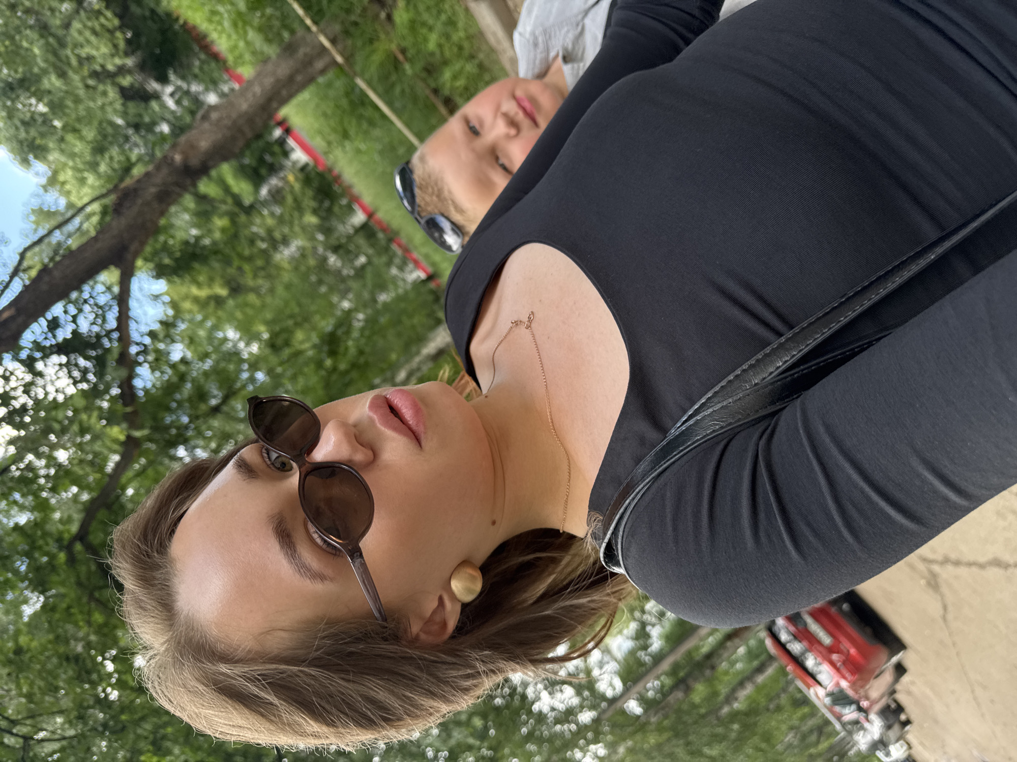 In the foreground, a young woman with light brown hair and sunglasses is taking a selfie. She is wearing a black long-sleeved shirt and large gold earrings. Her expression is neutral, and her lips are slightly parted. A thin gold necklace is visible around her neck. To her right and slightly behind her, a young boy with blonde hair and sunglasses perched on his head looks towards the camera. He is wearing a light-colored shirt. The background is blurred, showing trees, greenery, and a hint of a sky. To the left of the frame, parked cars, including a red one, are visible on a street or parking area. The scene appears to be outdoors, likely in a park or residential area, possibly in the city of Chaikovsky, Russia, given the context.