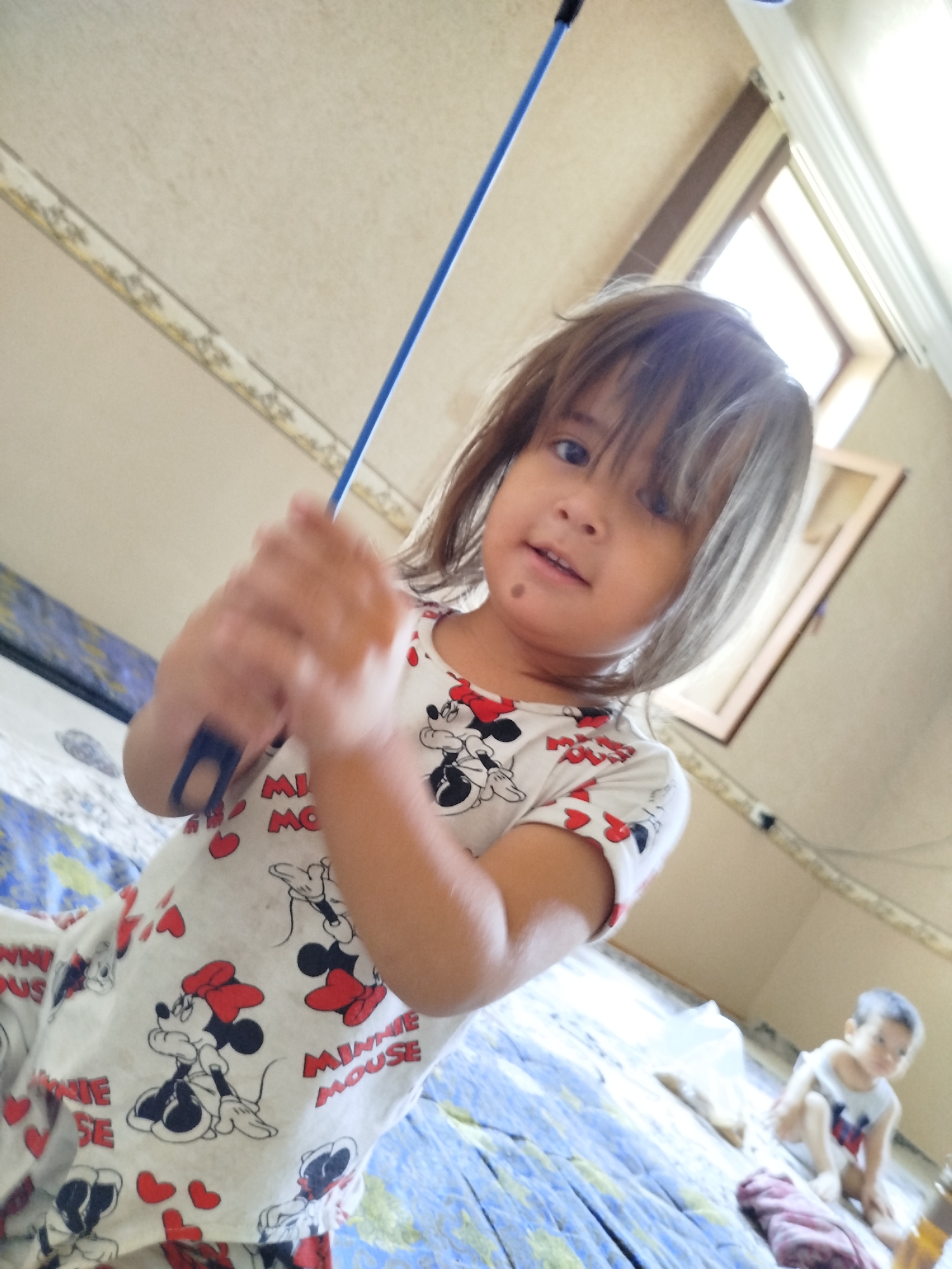 In Kokand City, Uzbekistan, a young girl with brown hair is captured in a low-angle shot, holding a blue pole. She is wearing a white t-shirt adorned with Minnie Mouse graphics. Her left arm, with a dark rectangular marking, is extended towards the camera. In the background, another young child, wearing a white shirt with red and blue accents, is seated on a bed. The environment appears to be indoors, with a patterned ceiling and a framed object on the wall. The lighting suggests natural light entering from a window.