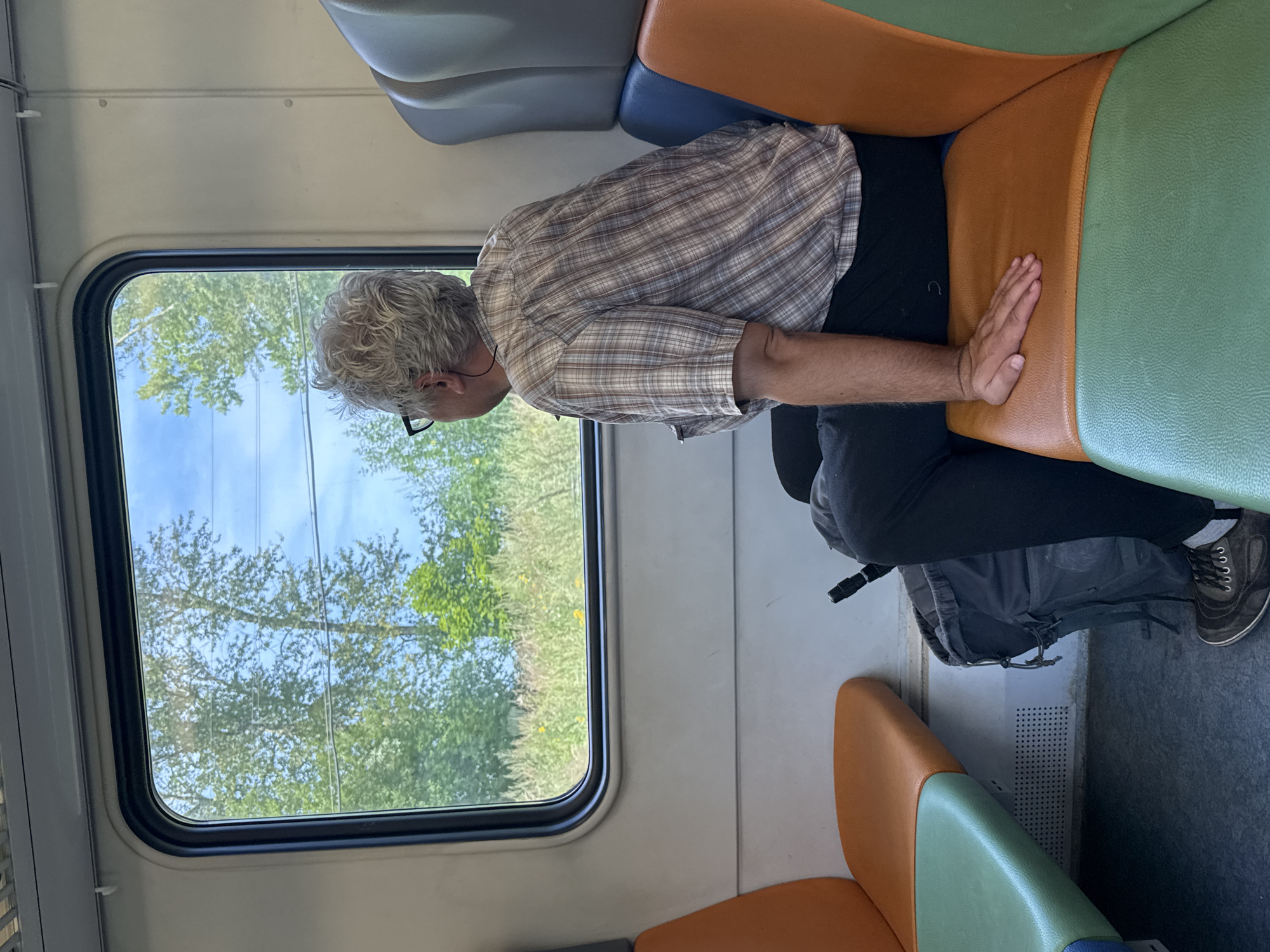 An adult male with grey, curly hair and glasses is seated on a train, looking out the window at a landscape of trees and grass under a blue sky. He is wearing a plaid shirt and dark pants. A grey backpack is on the floor beside him. The train interior features colorful seats in orange, blue, and green. The image was taken in Golitsyno, Russia.
