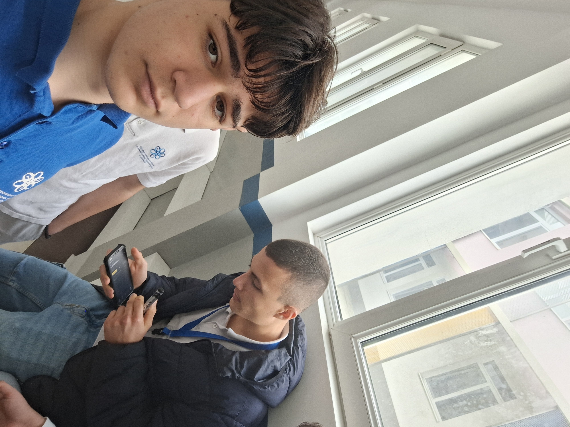 The image depicts two young men indoors, potentially in a building in Burgas, Bulgaria, given the flower logo seen on their shirts possibly relates to the flora in the city’s Sea Garden. One man, wearing a blue polo shirt with a flower logo, is positioned at the top of the frame. The second man, in a black jacket and white shirt, is holding a smartphone in his left hand and a pen in his right. He is seated and seems to be looking at the phone. A third person, only partially visible, is wearing a white polo with a flower logo similar to the first man. The setting is adjacent to a large window, with views of buildings outside. The windows are in the background.