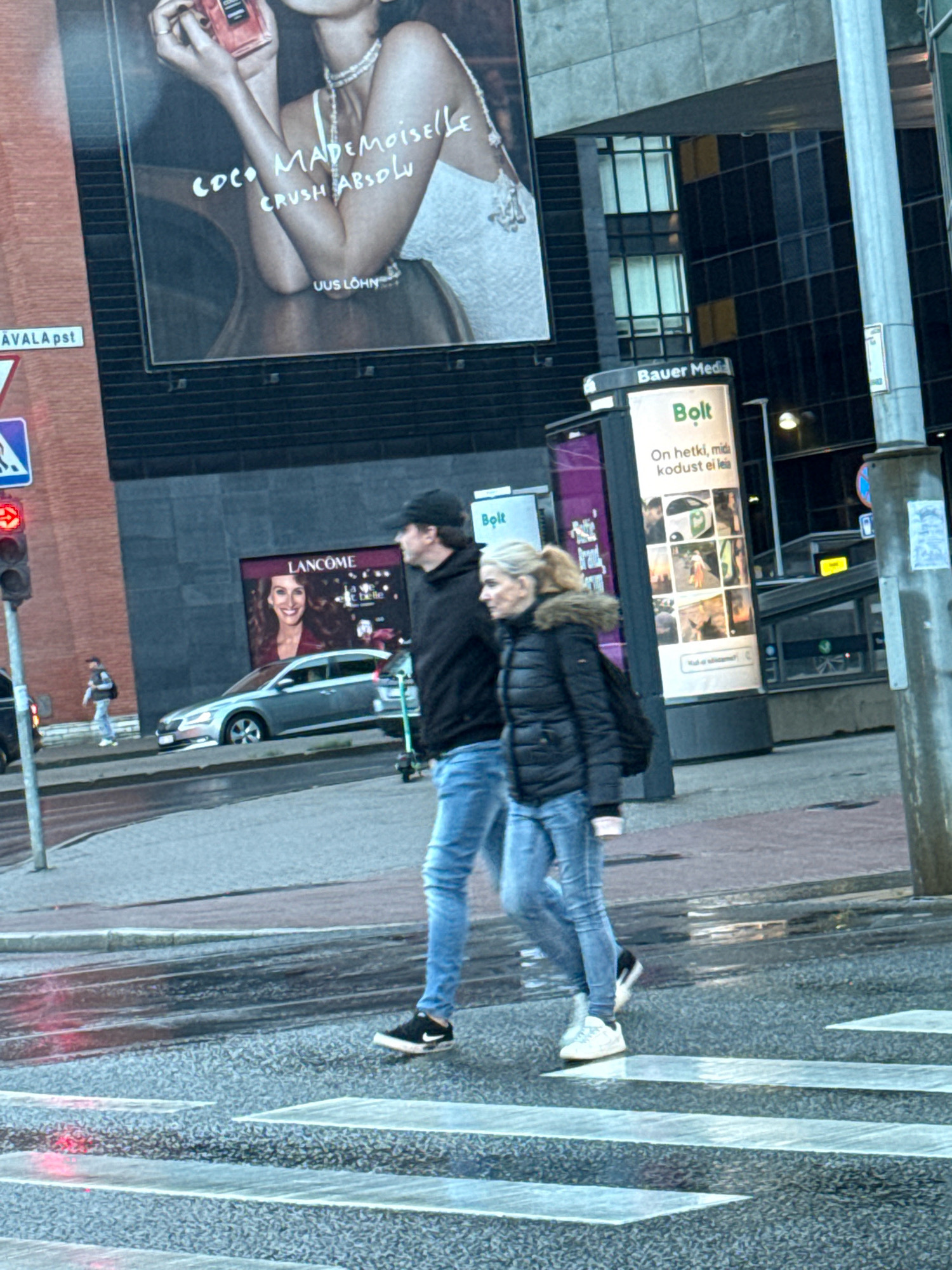 The image shows a street scene in Tallinn, Estonia, likely in the business district, given the modern architecture and advertising billboards. Two individuals, a man and a woman, are walking across a pedestrian crossing. The man is wearing a black hoodie, black baseball cap, and blue jeans. The woman is wearing a black puffer jacket with a fur-lined hood, blue jeans, and is carrying a black bag over her shoulder. They appear to be walking side-by-side.
In the background, several advertisements are visible. A large billboard features a black and white advertisement for "COCO MADEMOISELLE CRUSH ABSOLU" with text "UUS LÕHN" (New Fragrance). Below this, a smaller advertisement for "LANCOME" is displayed, featuring a smiling woman. Another prominent advertisement is for "Bolt," a ride-hailing service, with text "On hetki, mida kodust ei leia" (There are moments you can't find at home). The advertisements are mounted on modern buildings, one of which has a dark facade with checkered window patterns.
A silver car is parked on the street next to the Lancome advertisement. Traffic lights are visible, with the red light illuminated. A street sign with a blue triangle and white symbol is also present. The street surface is wet, suggesting recent rain. A lamppost and a utility pole are also part of the scene. The overall atmosphere is urban and commercial.