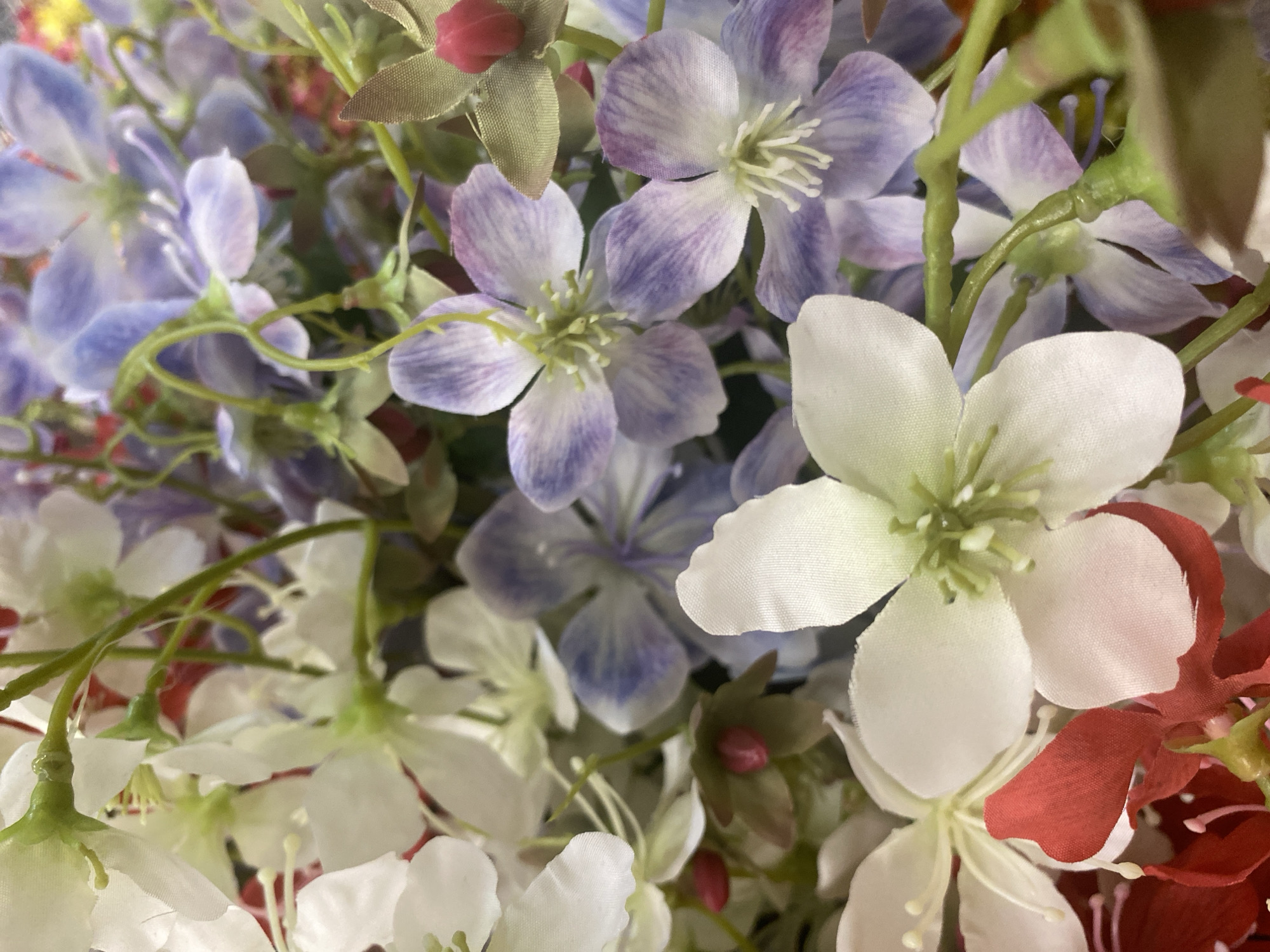 The image is a close-up, eye-level, full-frame shot of a bouquet of artificial flowers. The flowers are primarily white and lavender, with some red accents visible at the bottom. The arrangement appears to be a mixed bouquet, with various flower shapes and sizes. The lighting is natural and diffused, casting soft shadows. The background is a blur of more flowers, creating a shallow depth of field. The image does not provide any information or context about a specific location in Konya, Turkey, or any people or actions.