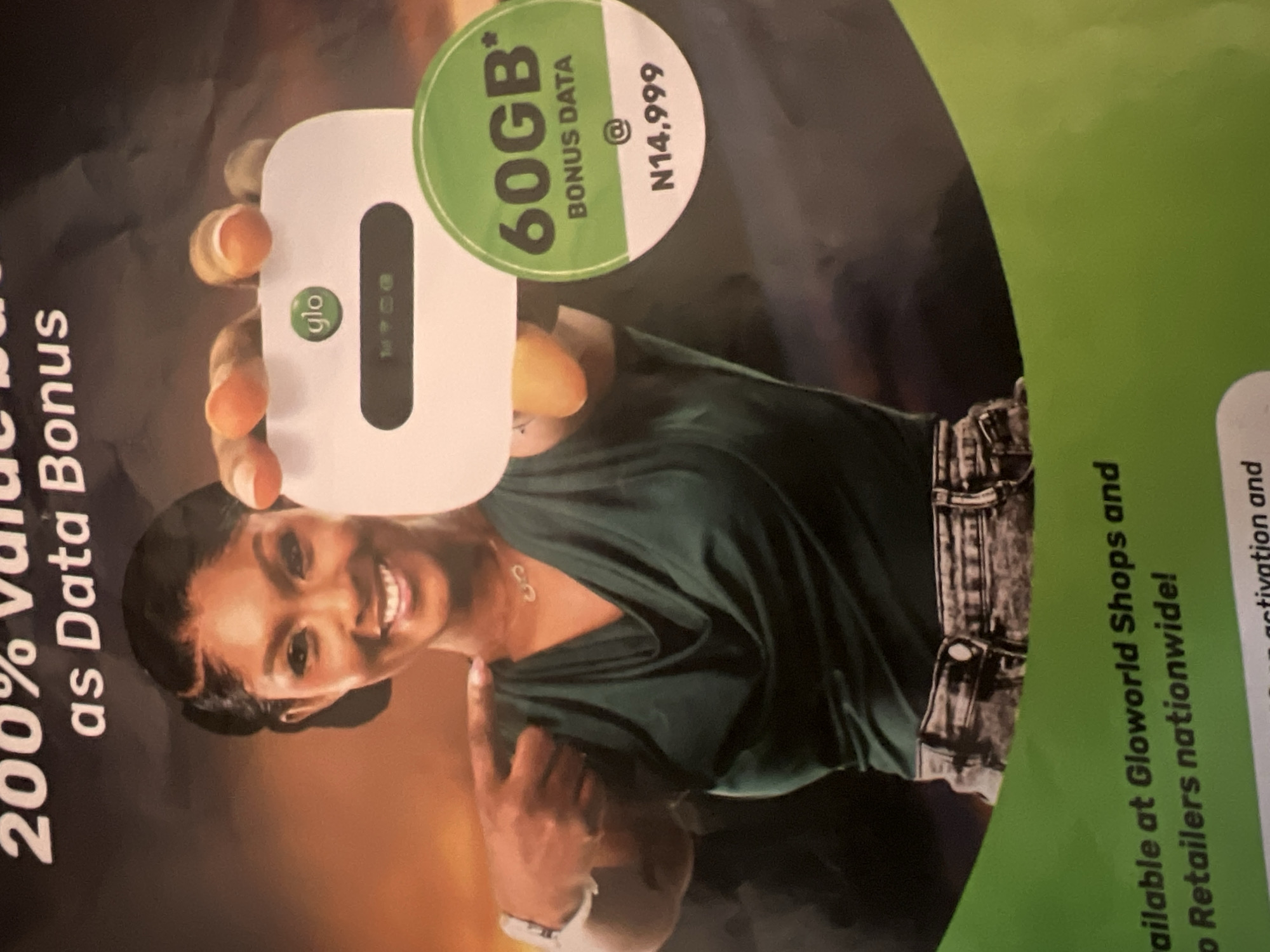 This is an image of an advertisement for Glo, a telecommunications company. In the foreground, a woman is smiling and pointing to a white device with the Glo logo. The device appears to be a mobile hotspot. Next to the device, a green circular sticker displays "60GB* BONUS DATA @ N14,999". The background is dark and blurry, with some text at the top of the image that appears to say "200% value back as Data Bonus". The bottom of the image shows text that reads "ailable at Gloworld Shops and Retailers nationwide!". There is no visible indication of the city of Jalingo, Nigeria within the image.